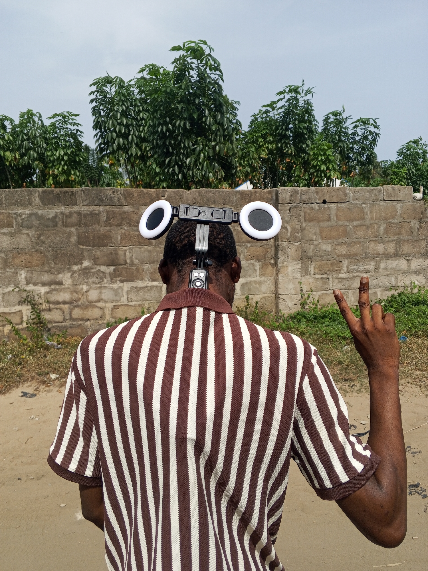 A person is seen from the back, standing in what appears to be an outdoor environment in Badagry, Nigeria. They are wearing a brown and white striped short-sleeved shirt and have a device attached to the back of their head, which includes two ring lights and a rectangular component. The person is holding up their hand, displaying a "peace" or "victory" sign. Behind the person is a low, textured brick wall, followed by a row of green trees. The ground is dirt or sand. The sky is visible in the background and is overcast.