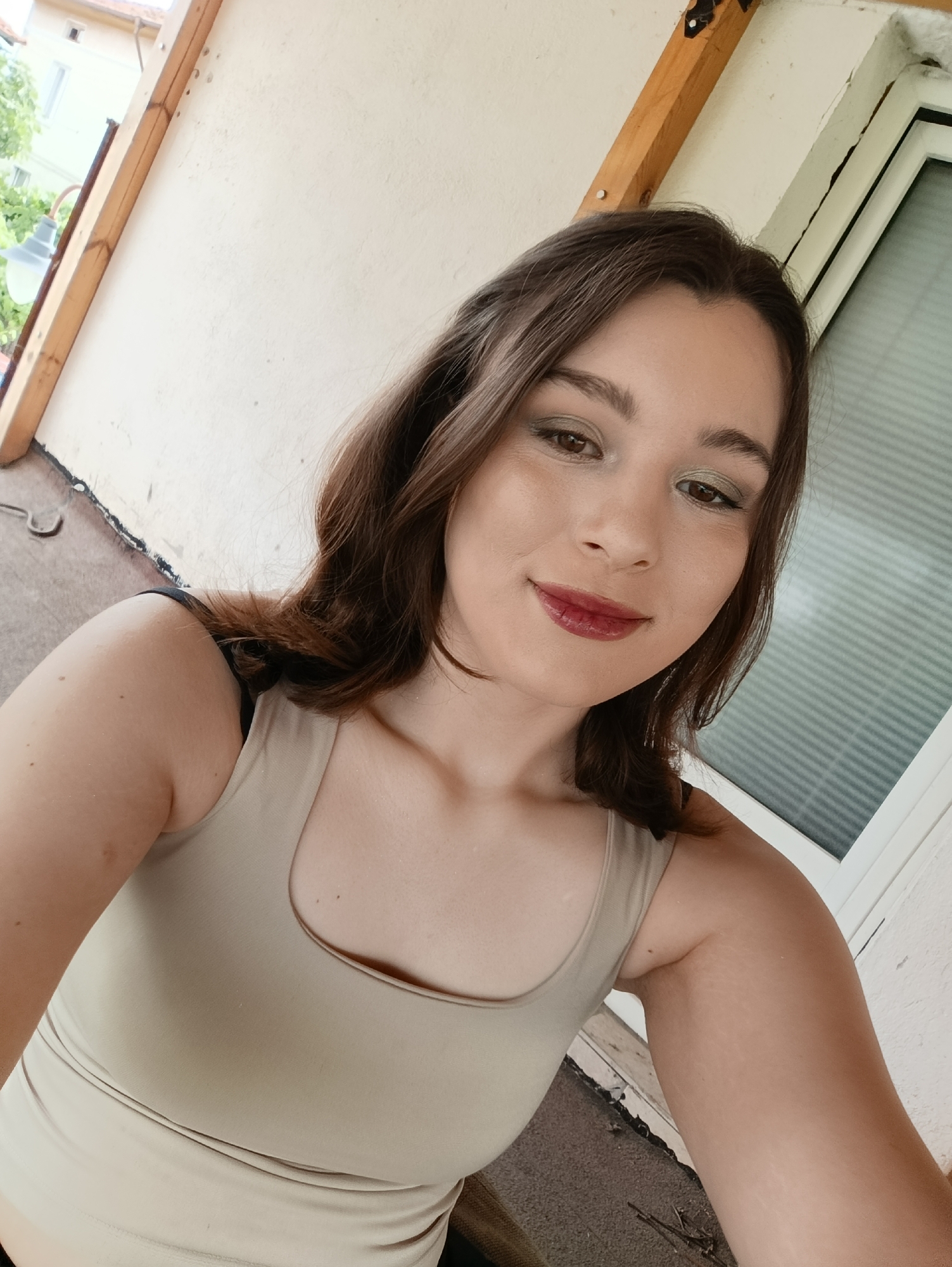 A young woman with brown hair and makeup is taking a selfie outdoors. She is wearing a beige tank top. The background shows a building with a white wall and wooden beams, a window with blinds, and some greenery. The scene appears to be on a balcony or porch.