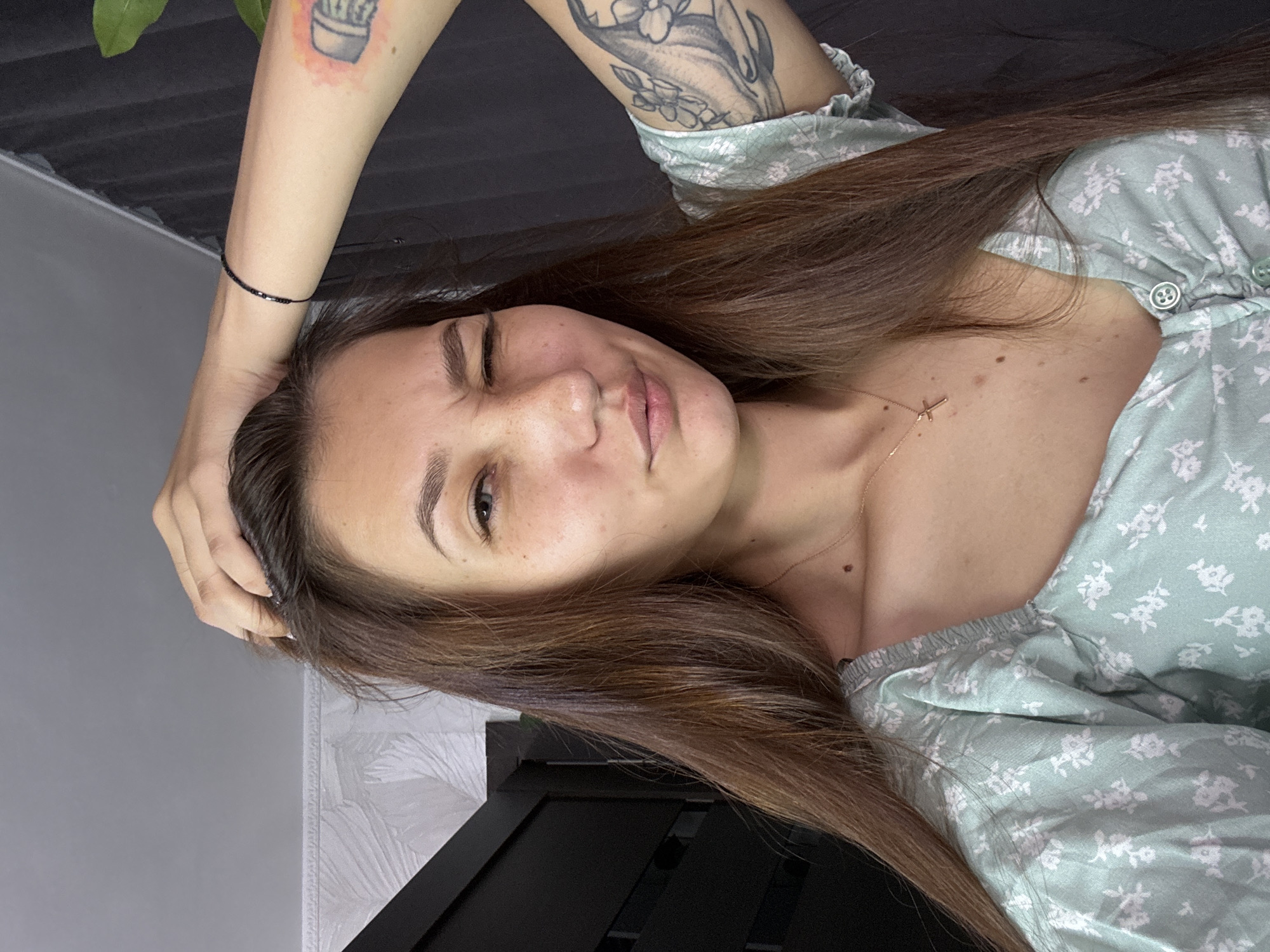 A young woman with long brown hair, wearing a light green floral top, is shown from the chest up. She is winking and has her hand on her head. She is also wearing a gold cross necklace and a black bracelet. Her arm has tattoos. The background is a dark curtain and a light-colored wall with a textured pattern. The location is not specified in the image.