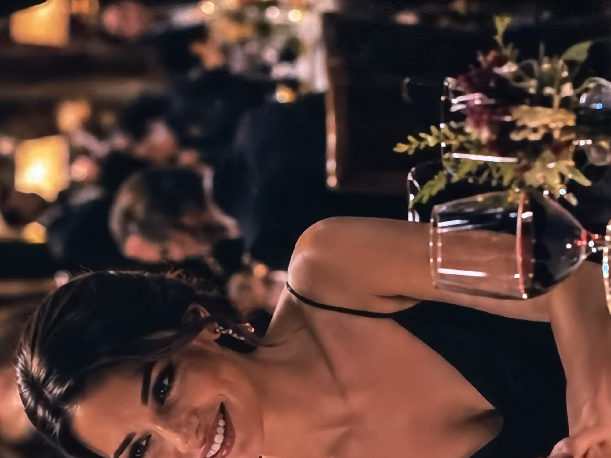 The image, originally rotated approximately 90 degrees counter-clockwise, captures an indoor formal event, likely an evening gathering or dinner. In the foreground, a young woman with dark hair and a warm smile is the primary subject, looking directly at the viewer. She is wearing a black, thin-strapped dress and has an earring visible on her right ear. Her expression suggests joy or contentment.
Behind her, the background is softly blurred, showing other individuals seated at tables, indicating a social event with multiple attendees. Warm, glowing lights, consistent with ambient or candle lighting, are visible in the background, reinforcing the perception of an evening setting.
On the table in front of or beside the woman, a wine glass filled with red wine is clearly visible. Nearby, a floral centerpiece, composed of green foliage and hints of red and yellow blooms, adds to the decorative elements of the event. The setting appears to be a sophisticated venue, such as a banquet hall or restaurant.
The activity taking place is a social gathering, with the woman possibly posing for a photograph or interacting with someone off-camera. Emotions conveyed by the woman are positive and cheerful. There is no visible text in the image. The location context is Sofiivska Borshchahivka, Ukraine.