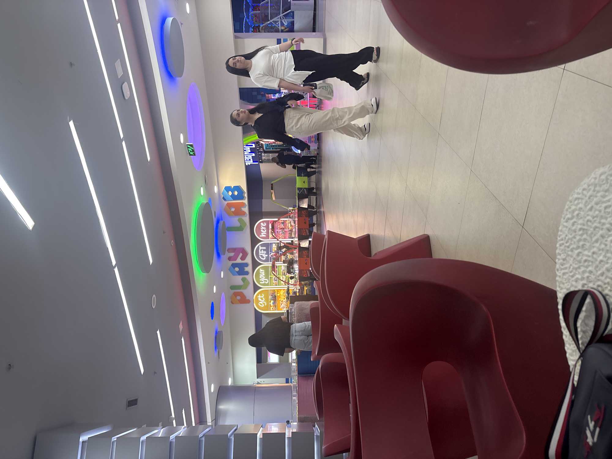 This image depicts an indoor scene within what appears to be an entertainment center or arcade, likely in Yerevan, Armenia.
In the midground, two young women are prominently featured. The woman on the left wears a black top and light-colored wide-leg pants, with white shoes. The woman on the right is dressed in a white top, black wide-leg pants, and black sandals, carrying a white bag. Both have long dark hair and appear to be walking or standing casually.
The foreground on the right side of the image contains several red, abstractly designed chairs and a partially visible black and white striped bag. The floor throughout the area is covered in light-colored tiles with dark grout lines.
In the background, an arcade area is visible, bustling with various game machines featuring flashing lights. A large, colorful sign spells out "PLAY LAB," and beneath it, smaller signs read "get your gift here." Another person, seen from behind in a dark top and light jeans, is standing near the arcade machines.
The ceiling is brightly illuminated by numerous fluorescent strip lights, and a green exit sign is visible. Colorful circular lights in blue, purple, and green adorn the wall, contributing to the vibrant atmosphere of the indoor space. The lighting is artificial and bright, typical of a commercial entertainment venue.