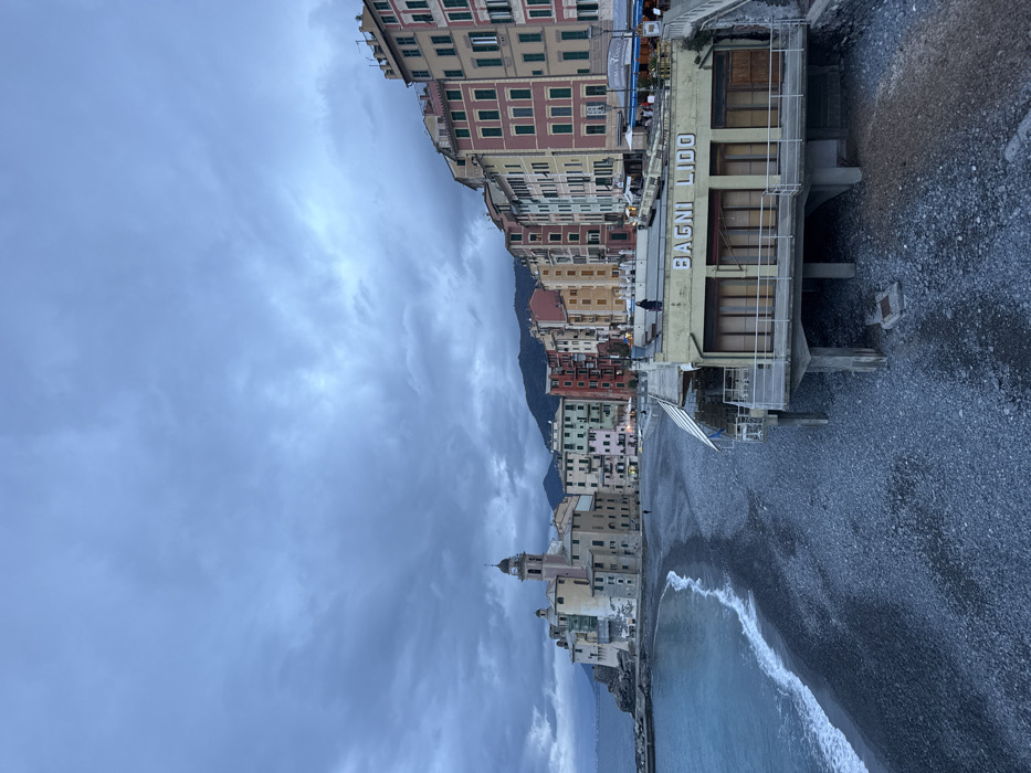 The image depicts the coastal town of Camogli, Italy, under an overcast sky. In the foreground, a dark pebble beach extends across the lower portion of the frame, with gentle waves breaking along the shoreline on the left. A two-story building, identified by the large white letters "BAGNI LIDO" on its facade, stands partially over the beach on concrete stilts, featuring multiple closed window/door panels on its ground level. A single individual, dressed in dark clothing, stands on the rooftop terrace of the "BAGNI LIDO" building, looking towards the right. An elevated walkway with stairs connects to the right side of this structure.
In the midground, a prominent row of tall, narrow, multi-colored buildings, ranging from pinks, reds, and yellows to greens and creams, lines the waterfront, ascending the hillside from left to right. Many windows feature dark green shutters. To the far left, built on a rocky promontory, stands a large, light-colored church with a domed roof and a clock tower, flanked by other pale-colored structures. A short breakwater extends into the sea from the church's base. Several faint lights are visible from businesses at the base of the colorful buildings. Two individuals are discernable on the pebble beach; one is walking near the waterline on the left.
The background features dark, forested hills rising behind the town. The sky is uniformly covered with thick, grey-blue clouds, indicative of overcast conditions or dusk. The sea, a deep blue, meets the cloudy horizon on the far left.