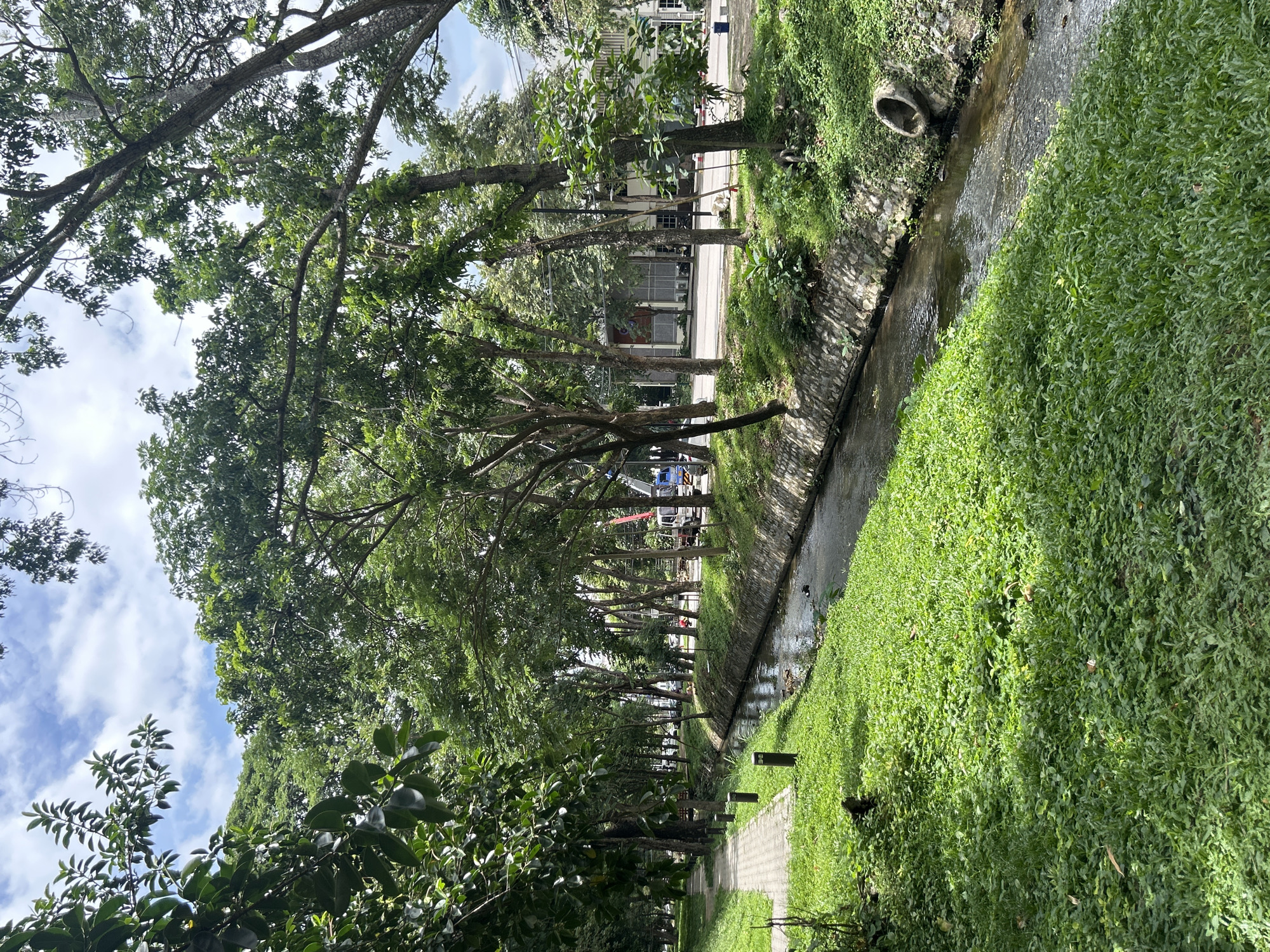 A narrow, concrete-lined canal flows through a park-like setting in Chiang Mai City Municipality, Thailand. The canal is bordered by lush green grass and lined with trees that cast dappled shadows. A paved walkway runs parallel to the canal on the left side of the image, marked by a series of black bollards. In the distance, a road with vehicles and construction equipment is visible. The sky above is partly cloudy with a mix of blue sky and white clouds. The scene conveys a peaceful urban environment with a focus on green spaces and water features.