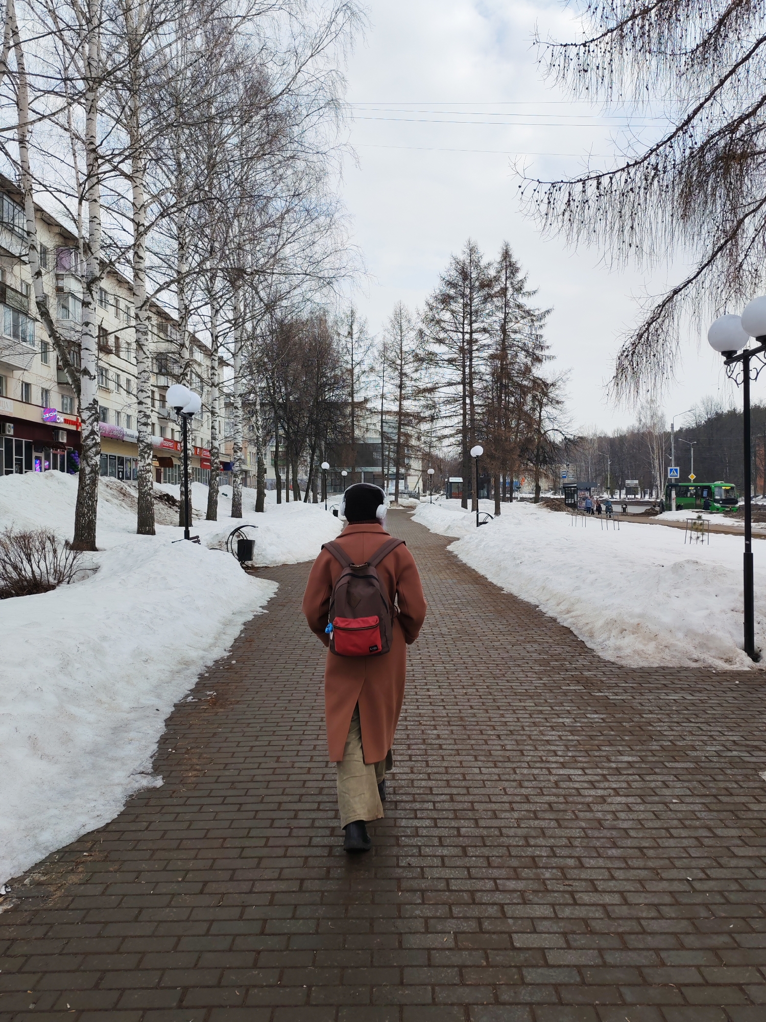 The image depicts a winter or early spring street scene in Chaikovsky, Russia. A single individual walks away from the camera on a wet, dark brown paved brick walkway that extends into the distance. The person wears a long, reddish-brown coat, light-colored trousers, black shoes, and white over-ear headphones, with a two-toned brown and red backpack visible on their back, from which a small blue item hangs.
Snowdrifts line both sides of the paved path, partially covering the ground and the bases of trees. To the left of the path, a row of tall birch trees with white bark and bare branches stands in front of a multi-story building featuring multiple windows and balconies. Ground-floor units of this building appear to be commercial establishments with illuminated signs, one of which reads "OZON." Several streetlights with spherical white lamps are positioned along the path.
To the right of the path, another row of trees, likely larch or similar conifers, with bare branches and small cones, separates the pedestrian area from a wider road. In the mid-ground, a green public transit bus is stopped at a bus stop, with several pedestrians visible near it. Distant vehicles and urban infrastructure are discernible beyond the bus stop. A wooded hillside rises in the far background. The sky is overcast and light grey.