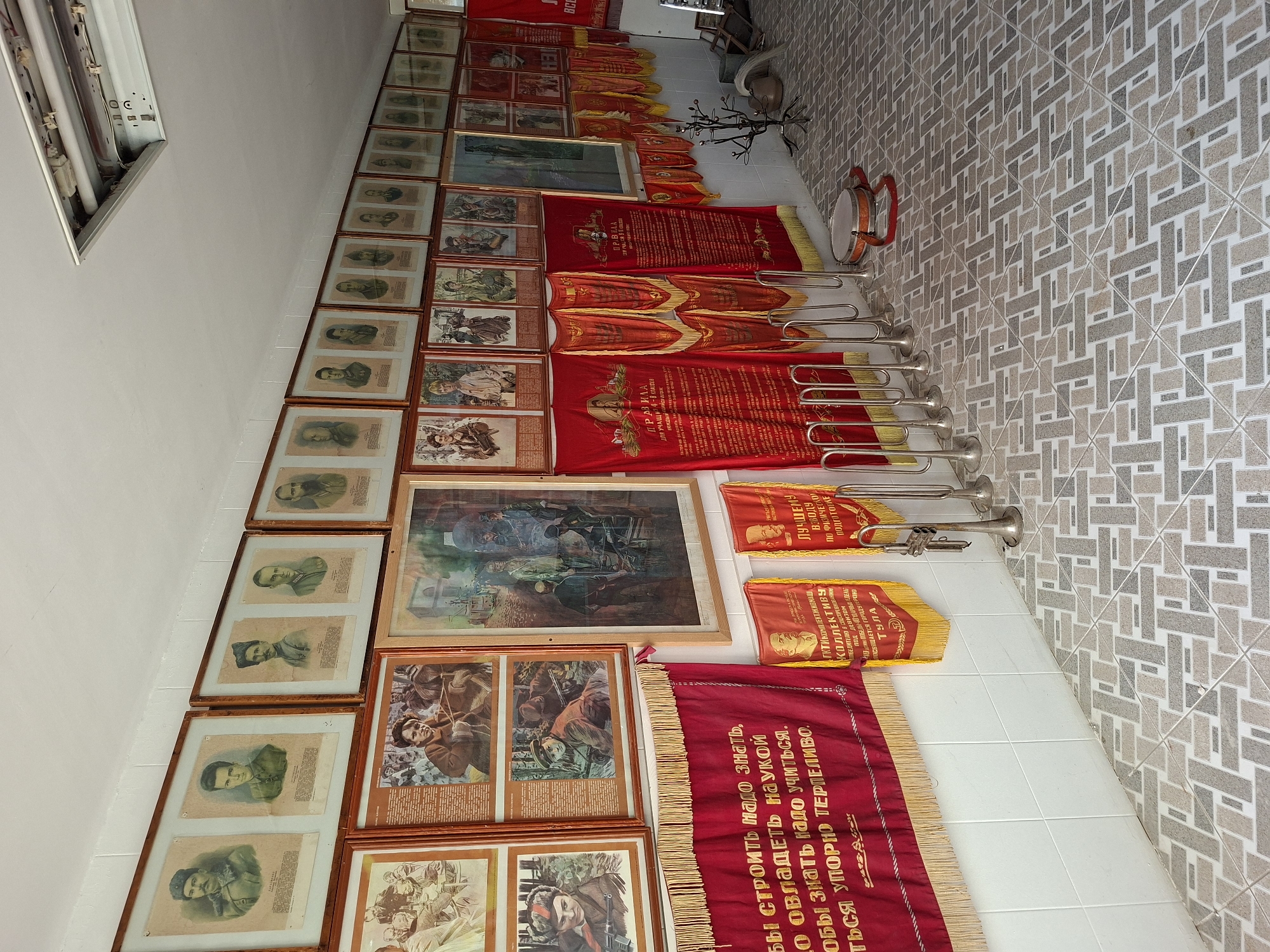 The image depicts an interior hallway or room with white tiled walls and a decorative tiled floor. The walls are adorned with numerous framed portraits and red banners, suggesting a museum, memorial, or cultural exhibit. The framed items on the wall are primarily portraits of individuals, many in historical attire, arranged in rows. Below these frames, several large red banners with yellow fringing hang, some bearing inscriptions in Cyrillic script. To the right, a row of brass instruments, possibly trumpets, stands upright. A small drum with a red strap is visible on the floor in front of the instruments, and a metal sculpture resembling a leafless tree is also present. Further down the hallway, more red banners are displayed, and the overall impression is of a space dedicated to historical commemoration or education.
This scene is located in the Cultural Center of Ialoveni, Moldova, which houses the Museum of History and Ethnography. The collection displayed includes portraits of historical figures, particularly those significant to the region's past, and banners that likely represent Soviet-era symbolism or historical events. The presence of musical instruments and the overall presentation are consistent with exhibits found in such cultural institutions in Moldova.
