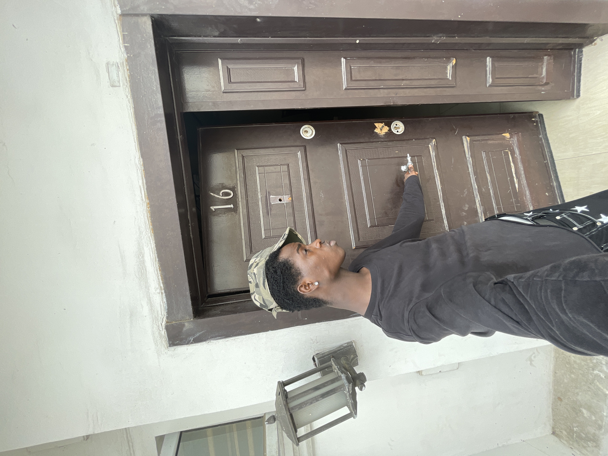A person is standing at a brown wooden door with the number 16 on it. The door is set within a brown frame, and the surrounding wall is light gray. The person is wearing a black long-sleeved shirt and a camouflage cap. A lantern-style light fixture is mounted on the wall to the side of the door. The location is Lagos, Nigeria.