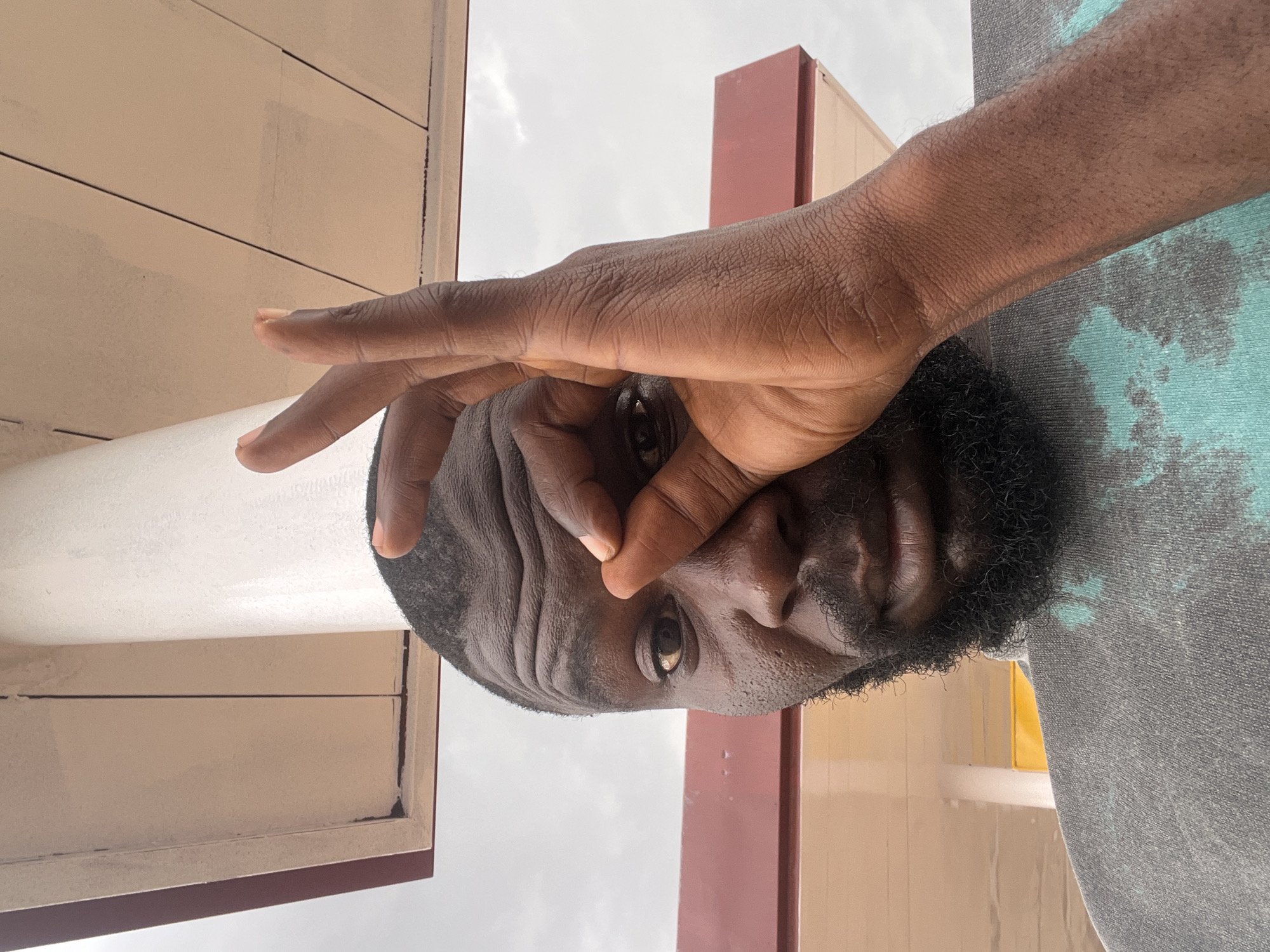 An outdoor portrait in Barakin Lerawi, Nigeria, features a man of color with a short beard and dark hair, looking directly at the camera. He is captured from a low angle, his head tilted slightly upwards. His right hand is raised, with his thumb and index finger forming a circle, and he peers through this opening. Wrinkles are visible on his forehead, and his expression appears neutral, possibly inquisitive or playful. He wears a grey t-shirt with a blue and black tie-dye pattern. The background consists of architectural elements: light beige or cream-colored panels with dark horizontal lines, and reddish-brown structural beams. The sky above is overcast, suggesting daytime with diffused light. No text is visible in the image.