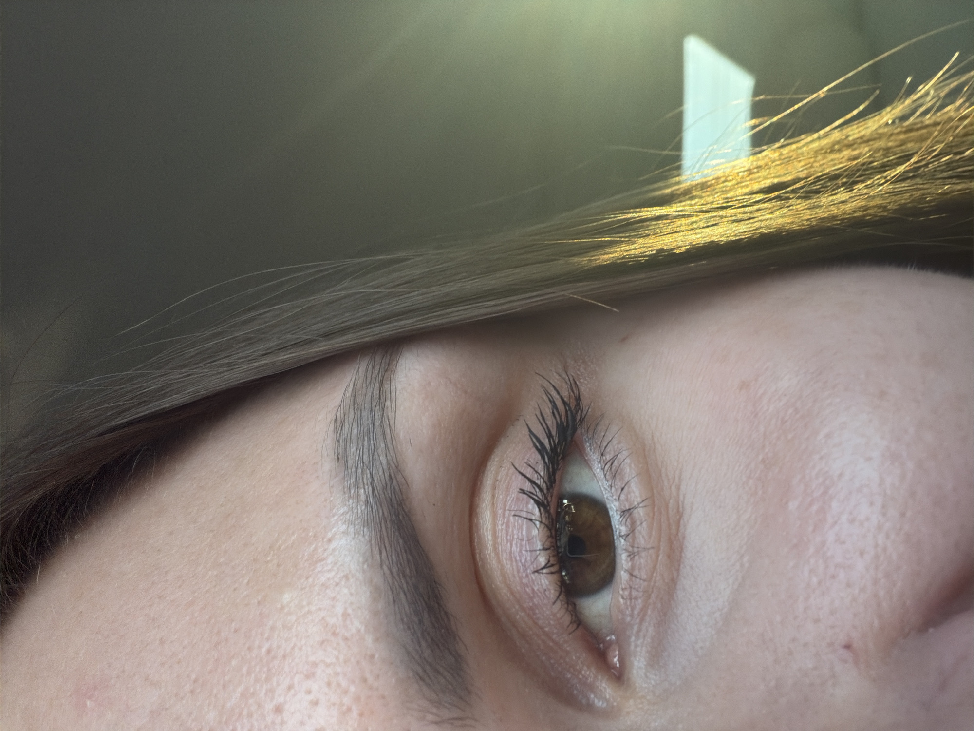 A close-up, eye-level, medium shot captures the right side of a person's face, focusing on the eye and forehead. The person has brown eyes and dark eyebrows, with fair skin that shows subtle pores and a few small blemishes. Light-colored eyeshadow is visible on the eyelid, and black mascara is applied to the eyelashes, which appear slightly clumped. The person's light brown hair cascades down the side of their face and shoulder, catching the sunlight which creates a warm, golden hue. The background is out of focus, showing a muted green-grey tone with a bright, rectangular light source visible on the right. The image appears to be taken indoors, possibly in a vehicle, given the quality of light and the presence of a reflective surface. The scene is forensic and lacks any specific identifiable markers to place it within Váhovce, Slovakia.
