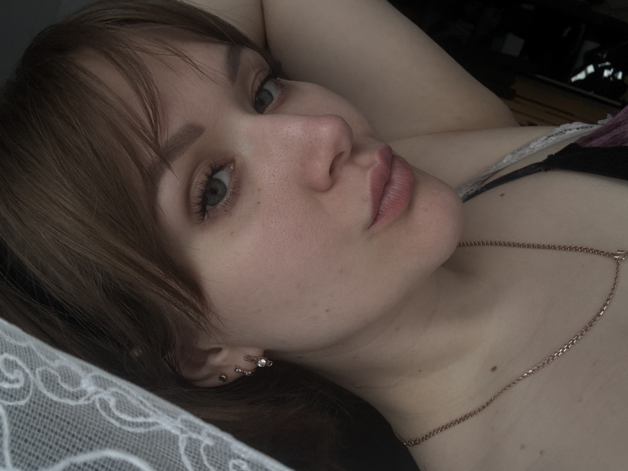 A close-up, angled shot depicts a woman with light skin, brown hair, and green eyes. She is lying down, with her head resting on a white lace pillow. A thin gold necklace is visible around her neck. She has piercings in her ear. Behind her, out of focus, is a dark space with wooden flooring. There is no information in the image that identifies Chaikovsky, Russia.