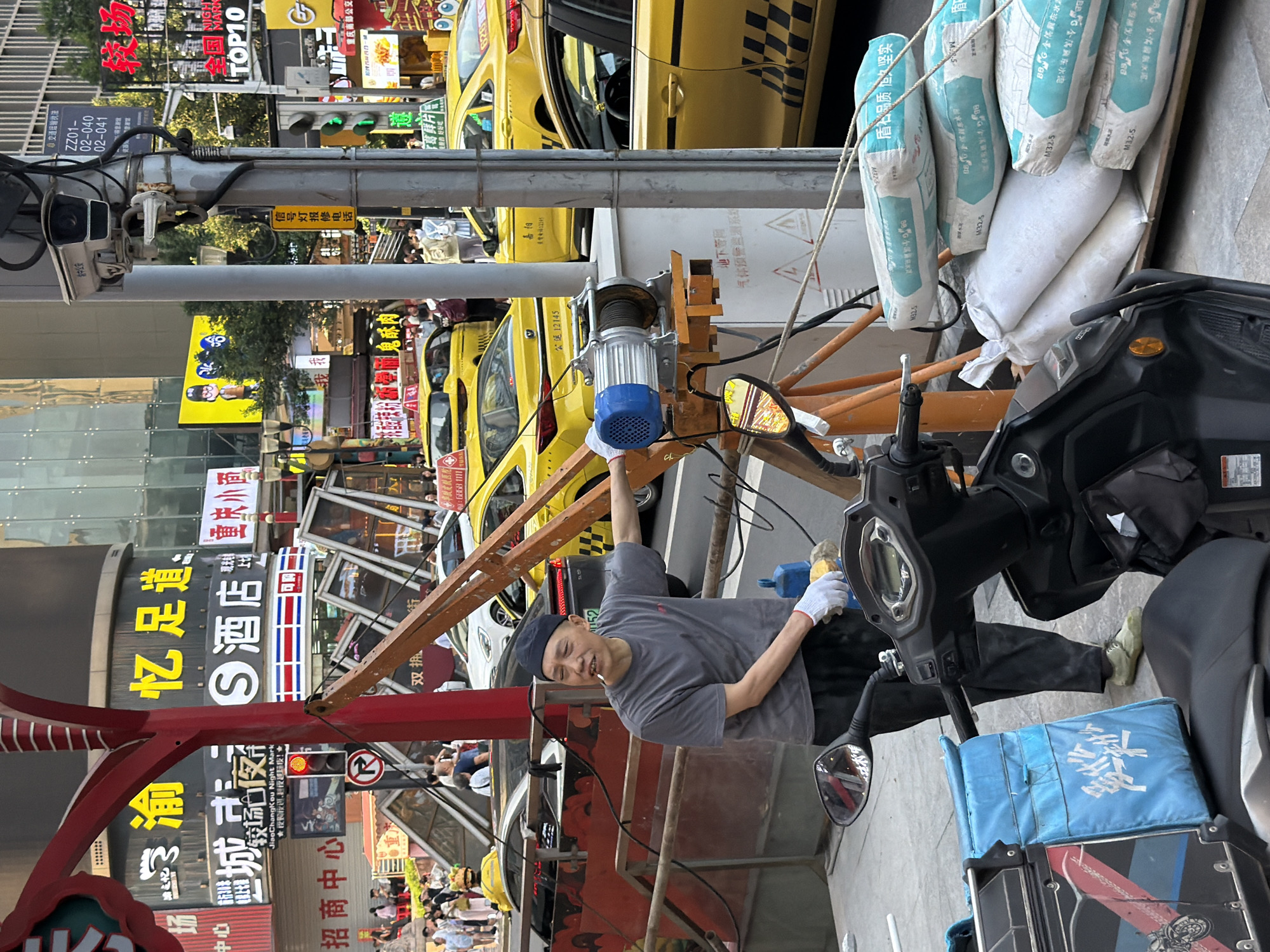 The image depicts a street scene in Yuzhong District, China. In the foreground, a man wearing a grey t-shirt, black pants, and a black beanie stands near a motorcycle. He is holding a cigarette in his mouth and wearing white gloves. In front of him, a blue delivery bag with Chinese characters is attached to the motorcycle. To the right of the man, a small crane with a blue electric winch is set up on a wooden platform. Bags of cement are stacked on a pallet to the right of the crane. In the background, several yellow taxis are parked, and a pedestrian walkway with people can be seen. Buildings with prominent signage, some in Chinese, line the street, suggesting a commercial area. The overall environment appears to be urban, with traffic signals and overhead wires visible.