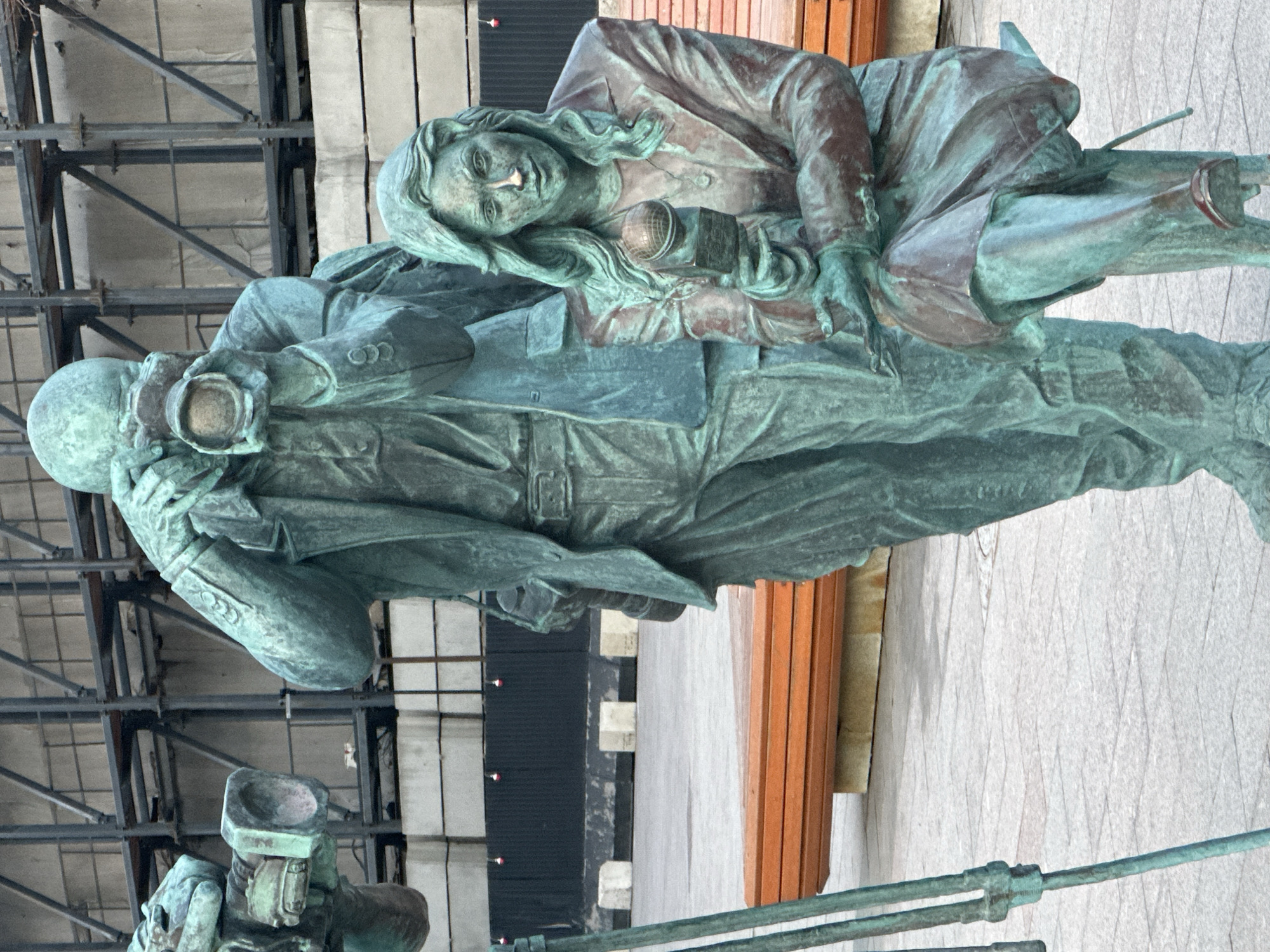 The image depicts a bronze statue, likely an outdoor monument, featuring two figures. One figure appears to be a prone male soldier or war correspondent wearing a helmet and holding a camera with a flash. The second figure, a female, is lying on top of him, holding a microphone.
The statue is positioned outside a modern building in Moscow, Russia. It is located in front of a wall and a wooden bench. The background shows structural elements of the building.