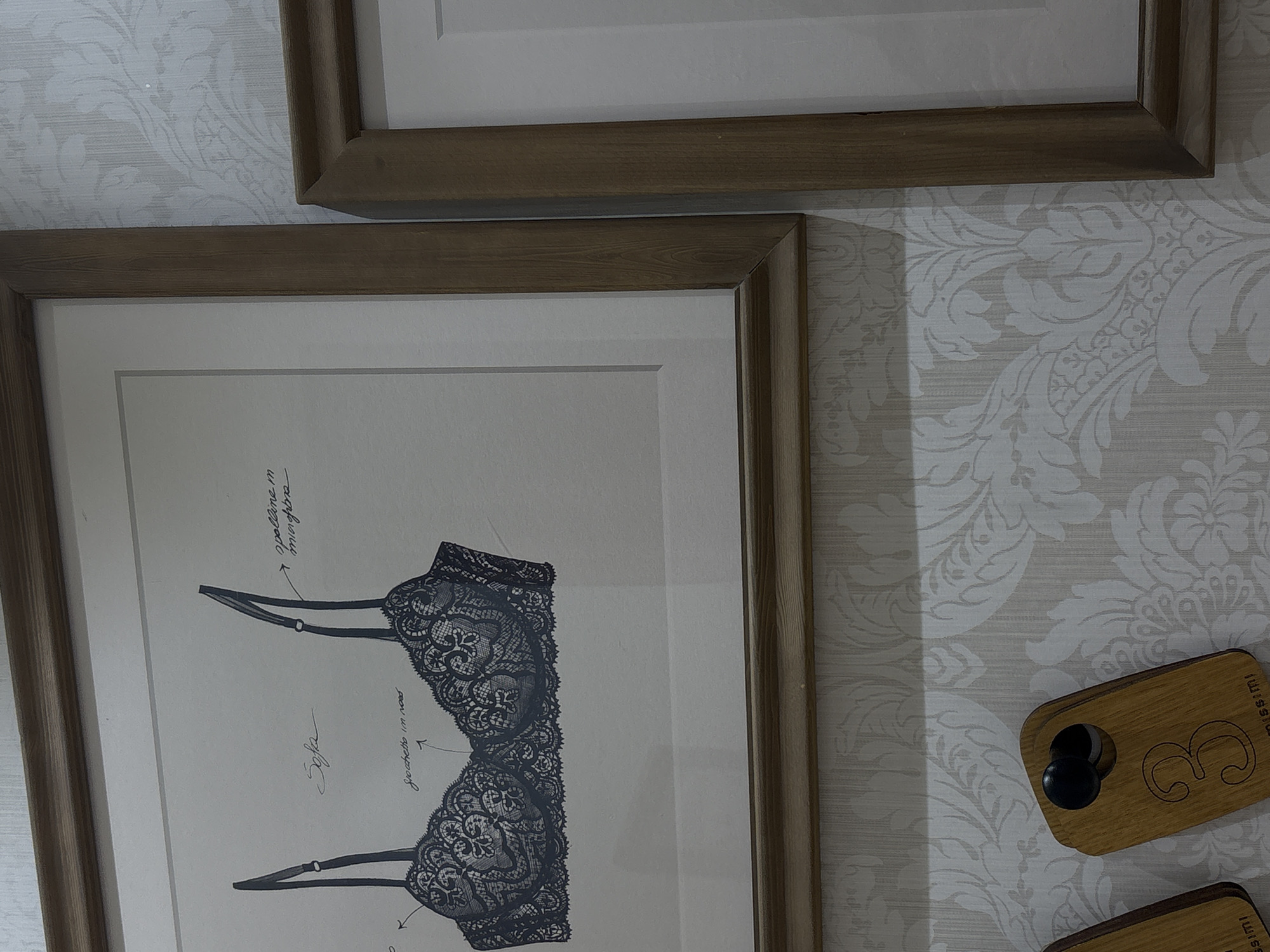 The image shows a close-up of two framed pictures hanging on a wall with patterned wallpaper. The left picture is a framed sketch of a lacy bra with handwritten annotations in Italian. The right picture is another framed artwork, partially visible, also with a white mat. Below the pictures, on the wallpaper, are two wooden tags shaped like keychains. One tag, clearly visible, is rectangular with rounded corners and has the number "3" carved into it, along with text that appears to read "missimi". The other tag, to the left, is partially visible and also seems to have text on it. The overall impression is that of a retail display, possibly in a boutique. The lighting suggests an indoor environment. There is no indication of the city of Chita, Russia, or any specific location information in the image itself.