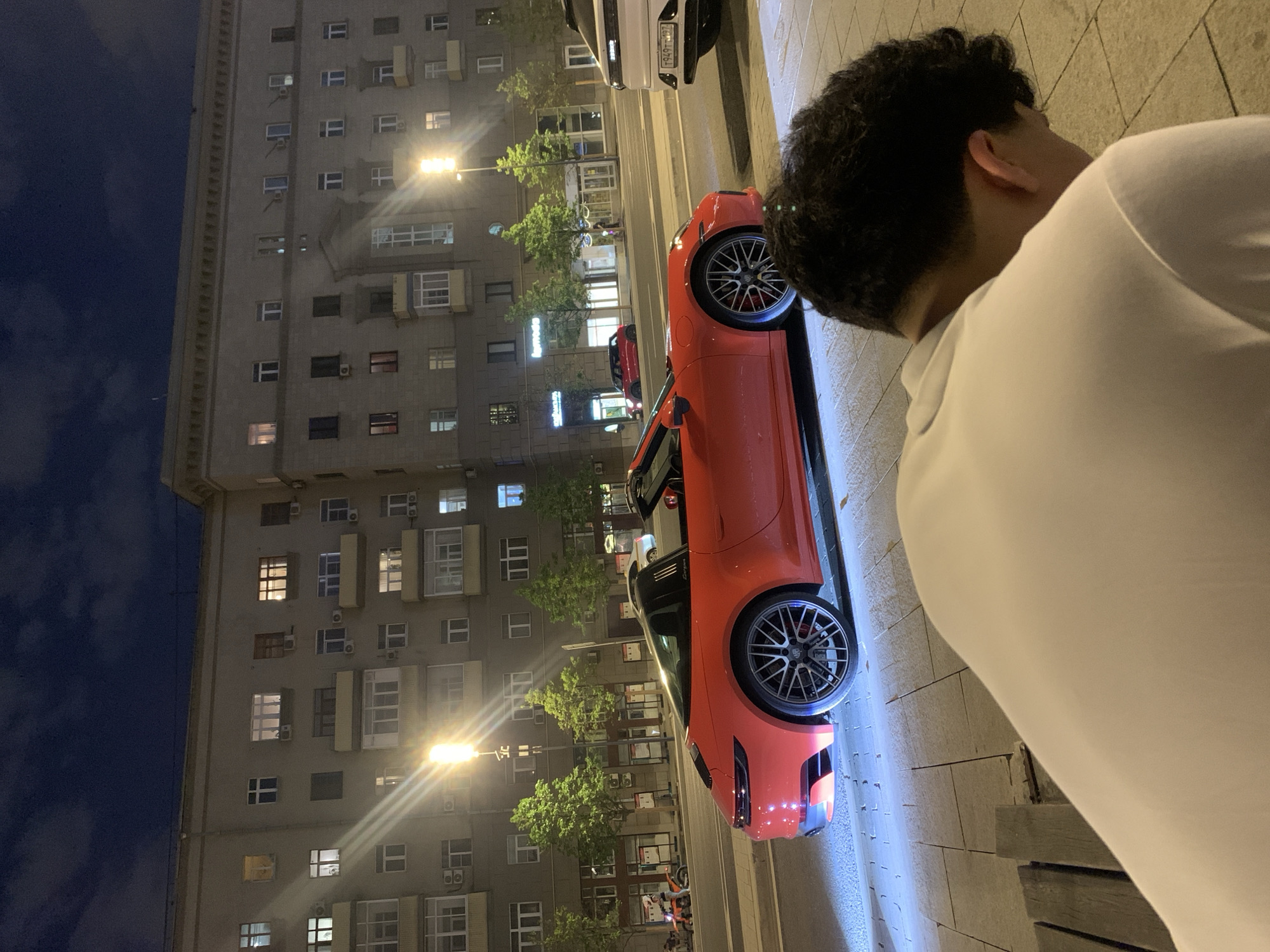 A nighttime urban street scene features a bright orange sports car parked near a tall, grey residential building. The building has multiple floors, each with numerous windows, and is illuminated by streetlights. Adjacent to the car, a person with dark hair and wearing a white shirt is partially visible. The street is paved with light-colored stones and appears wet. The scene is lit by streetlights, which reflect off the car and pavement, creating bright streaks of light. The sky is a deep blue, indicating it is night.