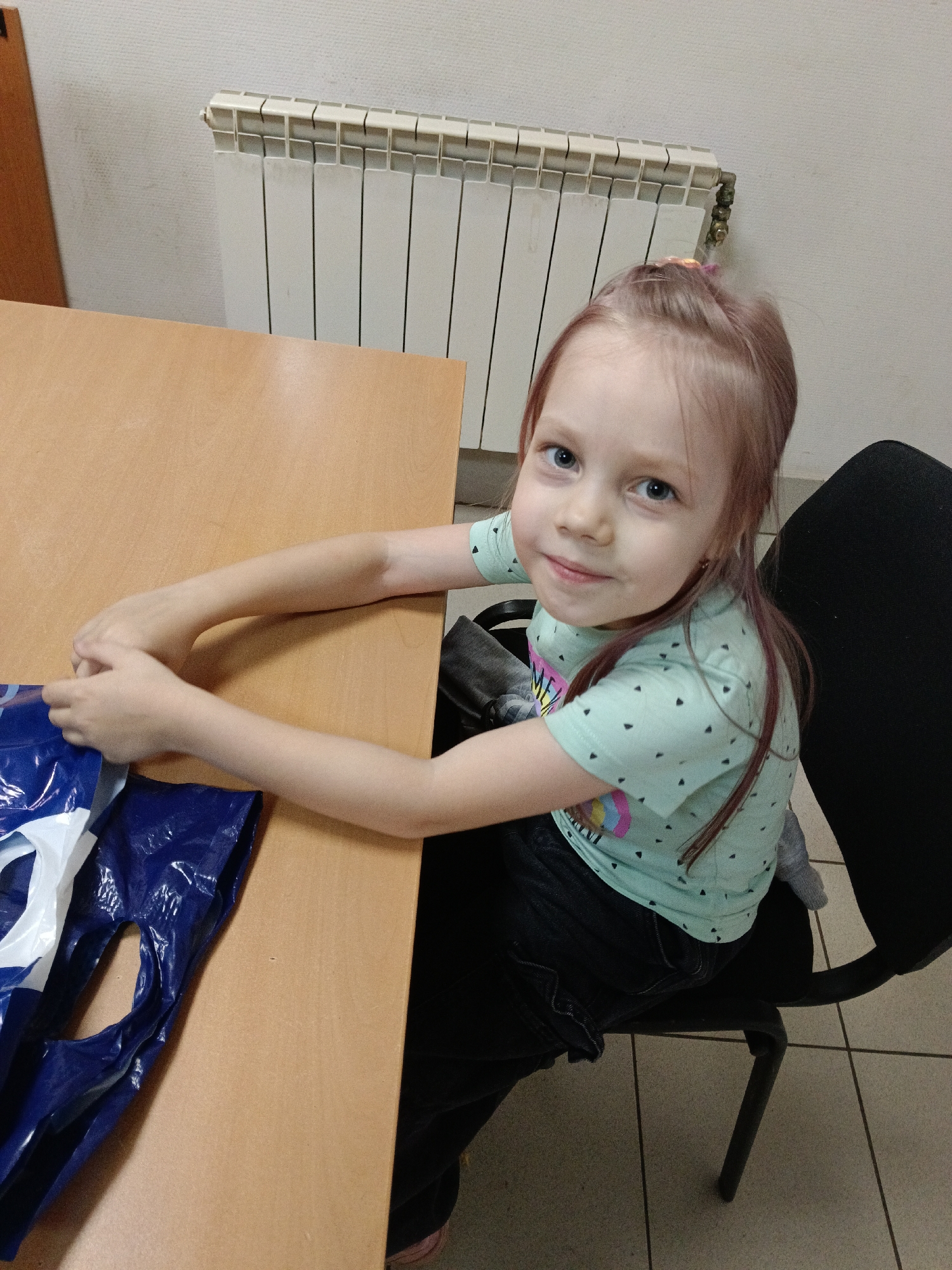A young girl with light brown hair pulled into a messy bun, tinged with pink, sits at a wooden table. She has blue eyes and is looking directly at the camera with a gentle smile. She is wearing a light blue t-shirt with small black triangle patterns and a colorful graphic on the front, along with dark denim jeans. Her arms are resting on the table, near a blue plastic bag with white accents. Behind her, a white radiator is mounted on a textured wall. She is seated in a black office chair with a metal base. The scene appears to be indoors, possibly in a classroom or office, given the radiator and the functional furniture. The lighting suggests it could be daytime, but there are no clear indicators of the time of day or weather conditions. The girl's expression is calm and friendly, suggesting a relaxed or perhaps slightly curious mood. There is no visible text in the image. The location context is Chaikovsky, Russia.