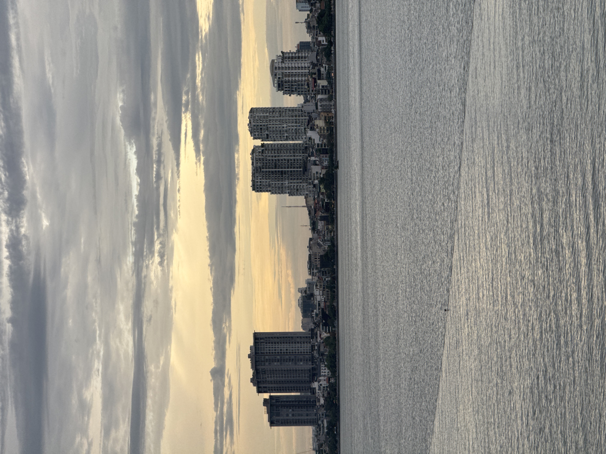 The image shows a view of the West Lake (Hồ Tây) in Hanoi, Vietnam, with a cityscape in the background. The sky is overcast with patches of sunlight breaking through, creating a soft, golden glow. The lake is calm with gentle ripples on its surface. In the middle of the lake, a single person is visible, appearing to be in a small boat or perhaps swimming. The city skyline features several modern high-rise buildings interspersed with older, lower-rise structures and lush green trees. The architectural style of the buildings suggests a contemporary urban environment. The overall atmosphere is serene and contemplative, characteristic of a cloudy day by a large body of water.