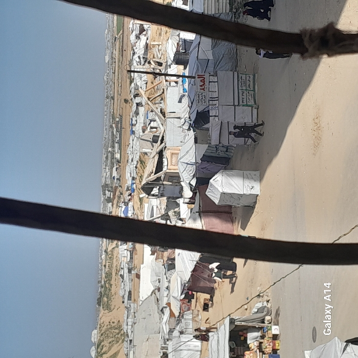 A shanty town or refugee camp sprawls under a bright sky, viewed through a window obscured by dark bars. Tents and makeshift structures crowd the sandy ground, showing signs of disrepair. Two figures in suits walk through the narrow pathways. Signs in Arabic adorn some buildings. The scene depicts poverty and displacement, contrasting with the implied safety of the viewer's location.