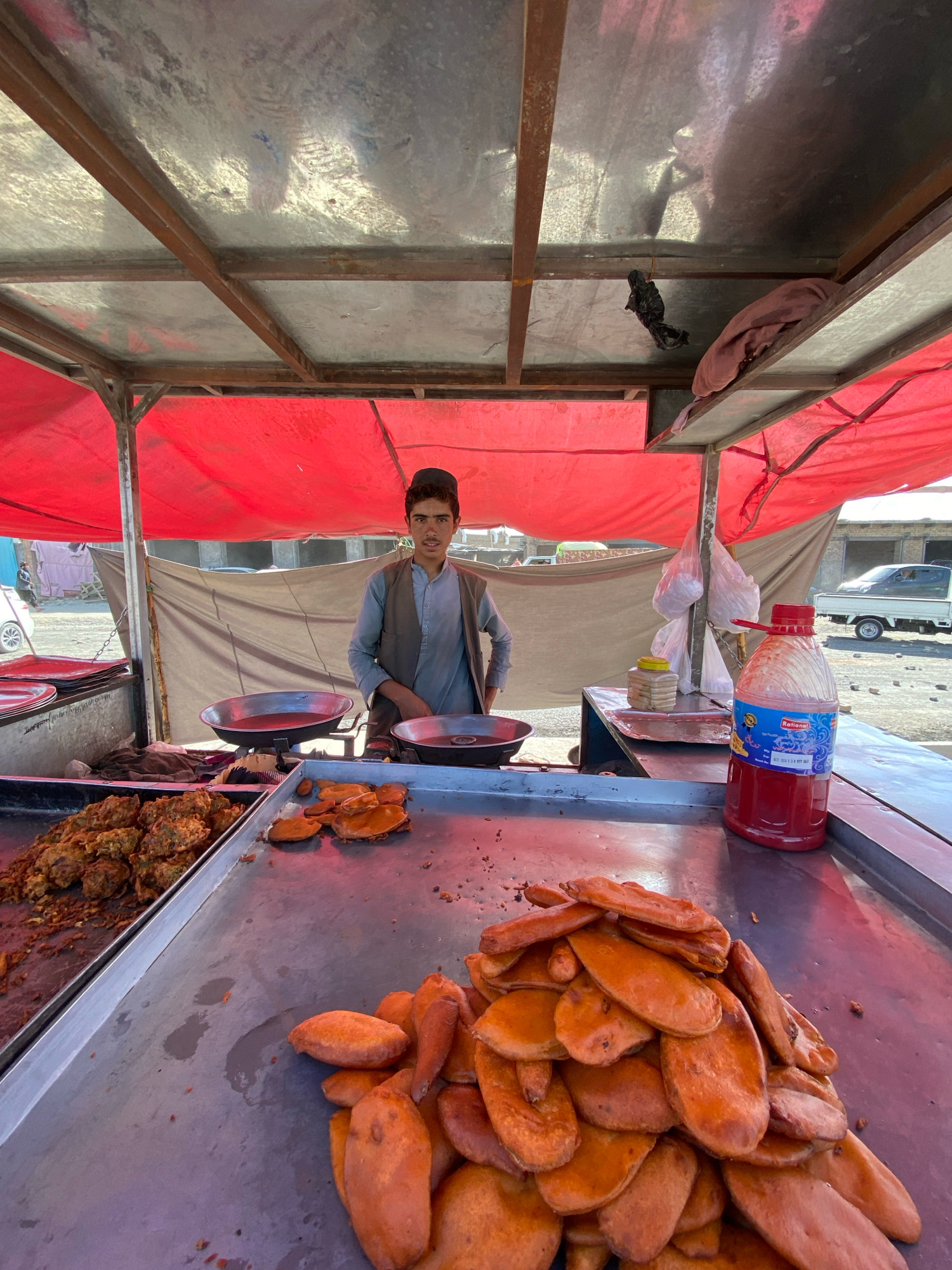 This image depicts a street food vendor in Spin Boldak, Afghanistan. A young man, wearing a grey vest over a light blue tunic and a black cap, stands behind a food stall. The stall is covered by a red tarp, and its metal roof is supported by rusted metal beams. In front of the vendor, a metal tray displays a pile of fried food items, which appear to be shaped like flat oval fritters. To the left, another tray holds a portion of fried food that resembles battered pieces of meat or vegetables. Two large metal pans, one containing what appears to be cooking oil, are set up on the stall. A bottle of red sauce, likely ketchup, with a label indicating it is "Rational" brand, sits on the counter to the right. In the background, a dusty street can be seen with a white car and a pickup truck parked. The environment suggests a marketplace or roadside area, typical of a bustling Afghan town.