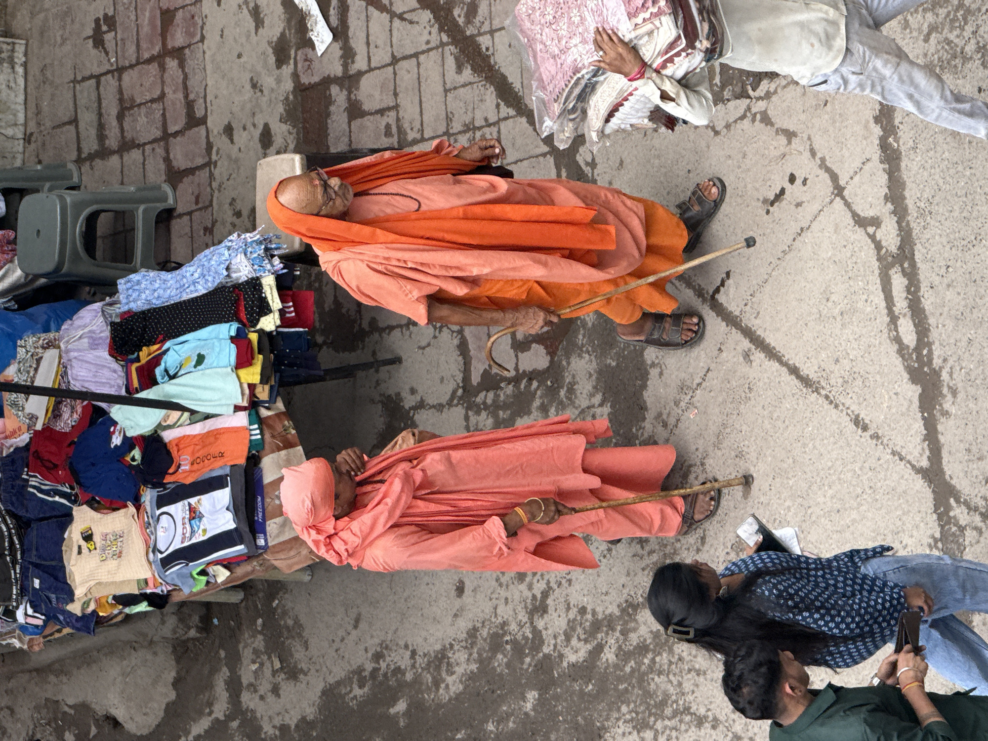 The image captures an elevated view of a street scene in Delhi, India, likely a market area. In the foreground, two individuals in saffron-colored robes are walking. The person on the left, wearing a head covering and a long robe with a scarf, leans on a wooden walking stick. The person on the right, an elderly man with glasses, also wears saffron robes and a beaded necklace, and walks with a similar walking stick.
To the left of these individuals, a young woman with long dark hair, wearing a blue printed top and jeans, holds a mobile phone. Beside her, a man in a dark green shirt, also in jeans, appears to be holding a wallet.
In the background, a stall displays a variety of colorful clothing, including t-shirts, shorts, and other garments. A gray plastic stool is visible near the stall. The ground is paved with brick-like tiles and concrete, with some water stains and debris scattered on the surface. To the far right, a person wearing a light-colored shirt and trousers carries a stack of folded fabric, possibly being transported from the market. The overall impression is a bustling, everyday street scene in a vibrant Indian city.