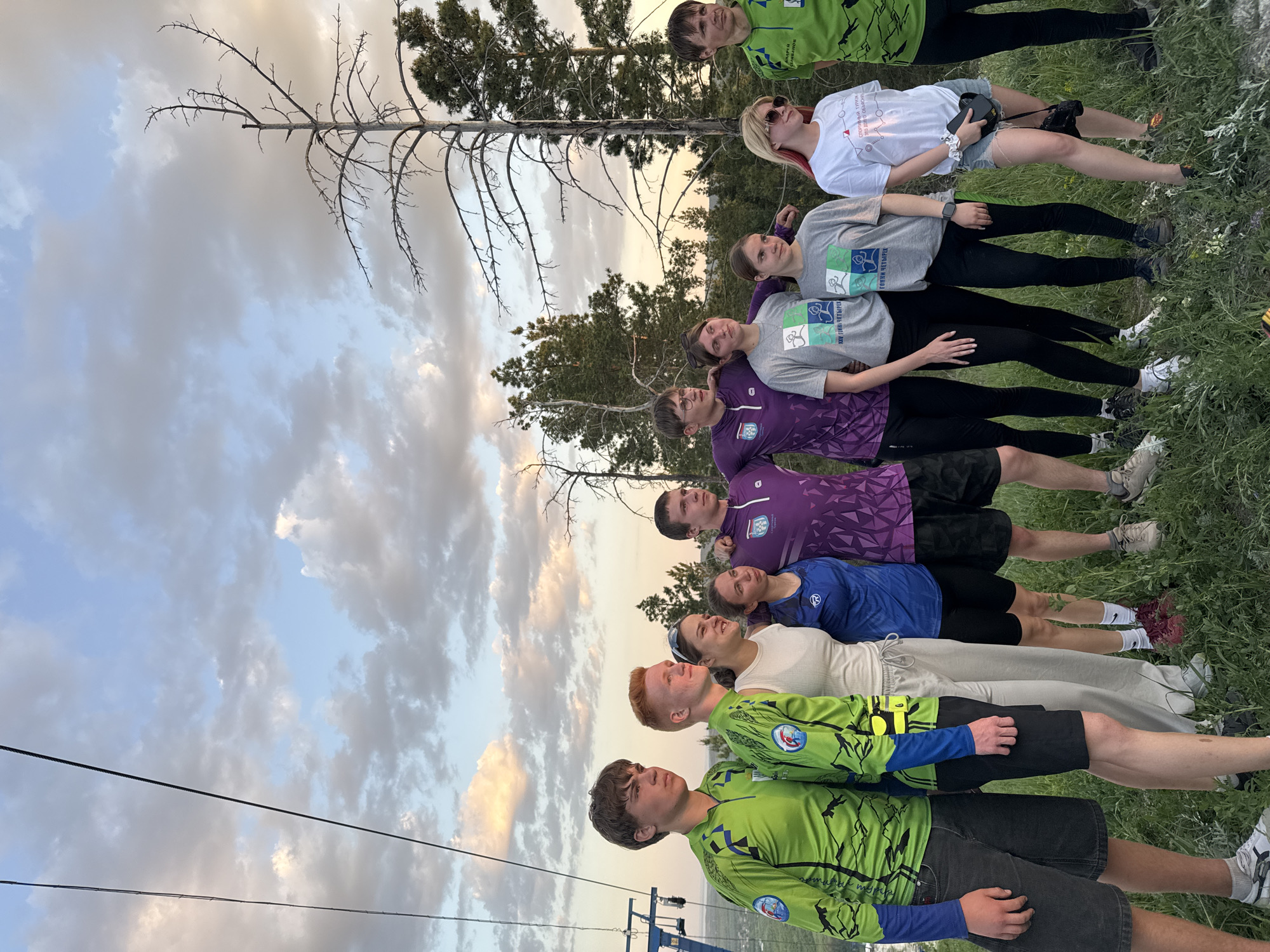 A group of ten individuals are lined up outdoors, seemingly on a grassy slope in Khvalynsk, Russia. The background includes trees, a partially cloudy sky, and a ski lift structure. The people wear different attire. At the bottom of the line, two men wear matching green and blue long-sleeved shirts with images of foliage, along with dark shorts. The next person wears a light-colored outfit, with a person following wearing blue. Two individuals wear purple shirts. Next are two women wearing gray t-shirts with an image on the front. At the top, a woman wears a white t-shirt and shorts, and another man wears green and blue.