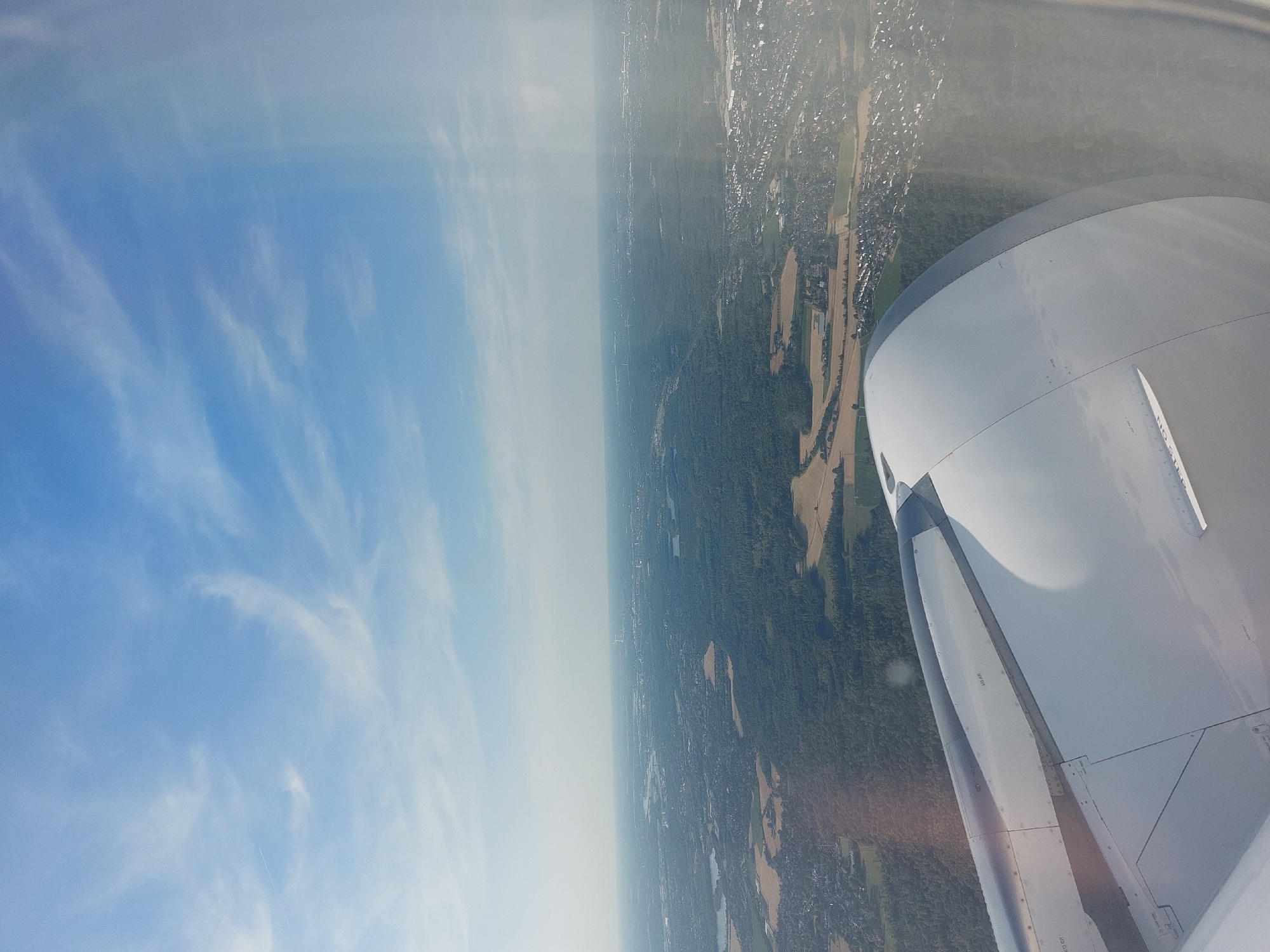 An aerial view from an airplane window shows a sprawling landscape of forests, fields, and urban development under a partly cloudy blue sky. The foreground is dominated by the white, rounded surface of an airplane engine nacelle and a portion of the wing, with visible panel lines and some faint text. Beyond the aircraft, the ground unfolds into a patchwork of dense green forests, lighter green fields, and scattered clusters of buildings. In the distance, a hazy cityscape stretches across the horizon, with what appear to be industrial facilities and a city skyline. The perspective suggests the aircraft is in descent or cruising at a moderate altitude over a densely populated region. Based on the landscape and visible urban structures, this scene is consistent with an overhead view of the city of Ratingen, Germany, and its surrounding areas.
