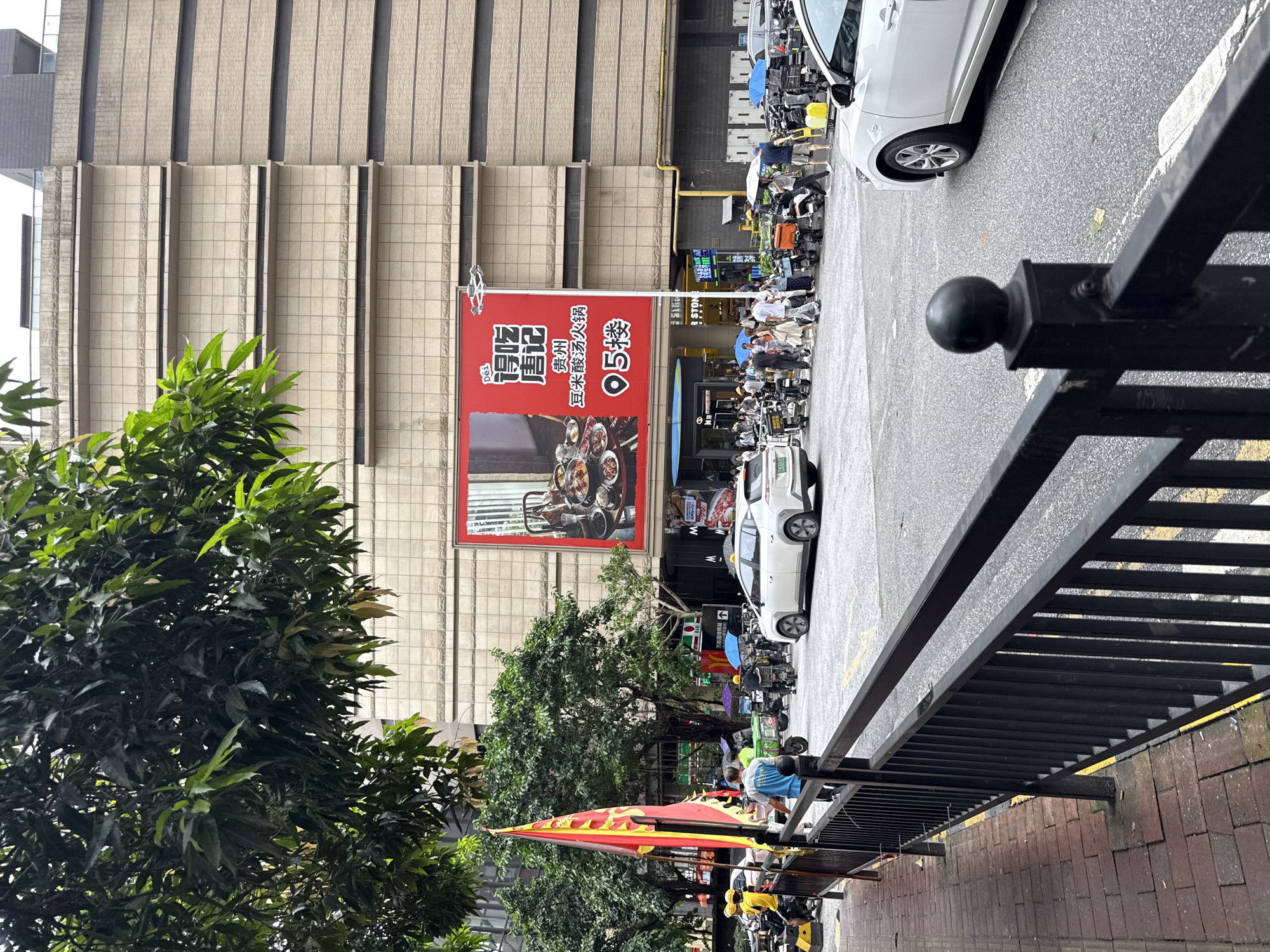 A street scene in Haizhu District, China, features a large building with a prominent red advertisement for "De Chi Tang Ji" (得吃唐记), a restaurant specializing in Guizhou sour soup hot pot, with its location indicated as "5th floor". In the foreground, a black metal fence runs along a brick sidewalk. The street is populated with various vehicles, including a white car, numerous motorcycles, and electric scooters, some of which are being ridden by individuals wearing yellow helmets, possibly delivery personnel. Several people are walking on the street, and some are holding umbrellas, suggesting it may be a cloudy or rainy day. Trees with lush green foliage are visible, with one large tree prominently positioned in the upper left portion of the frame. The building's facade is a light beige color, composed of tiled sections and horizontal bands.