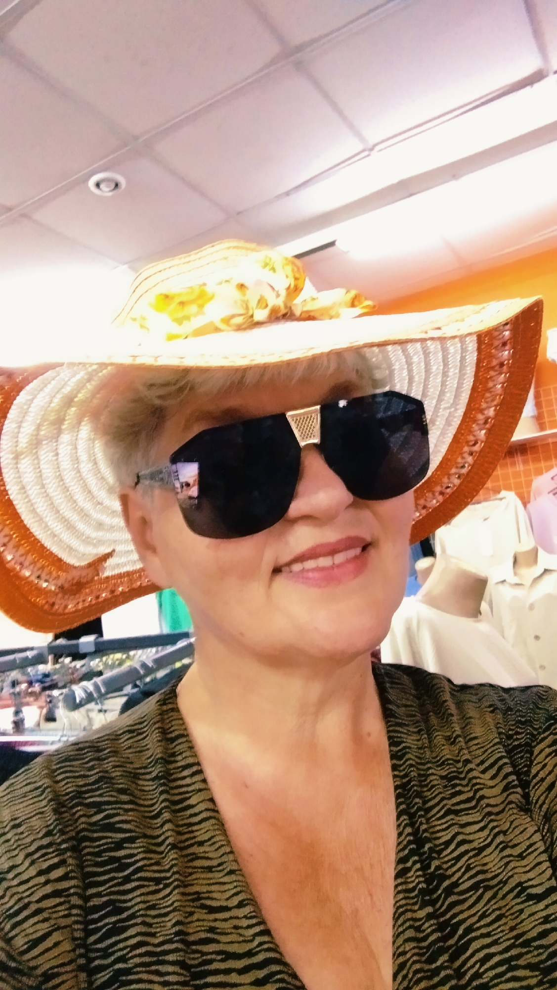 A woman wearing a wide-brimmed straw hat with a patterned ribbon and large, dark sunglasses poses for a photo. She has short, blonde hair and is smiling. She is wearing a dark, patterned top. The background appears to be an indoor setting, possibly a store, with clothing racks and mannequins visible. The ceiling lights are on. The image does not provide any specific information about the location being the city of Horishni Plavni, Ukraine.