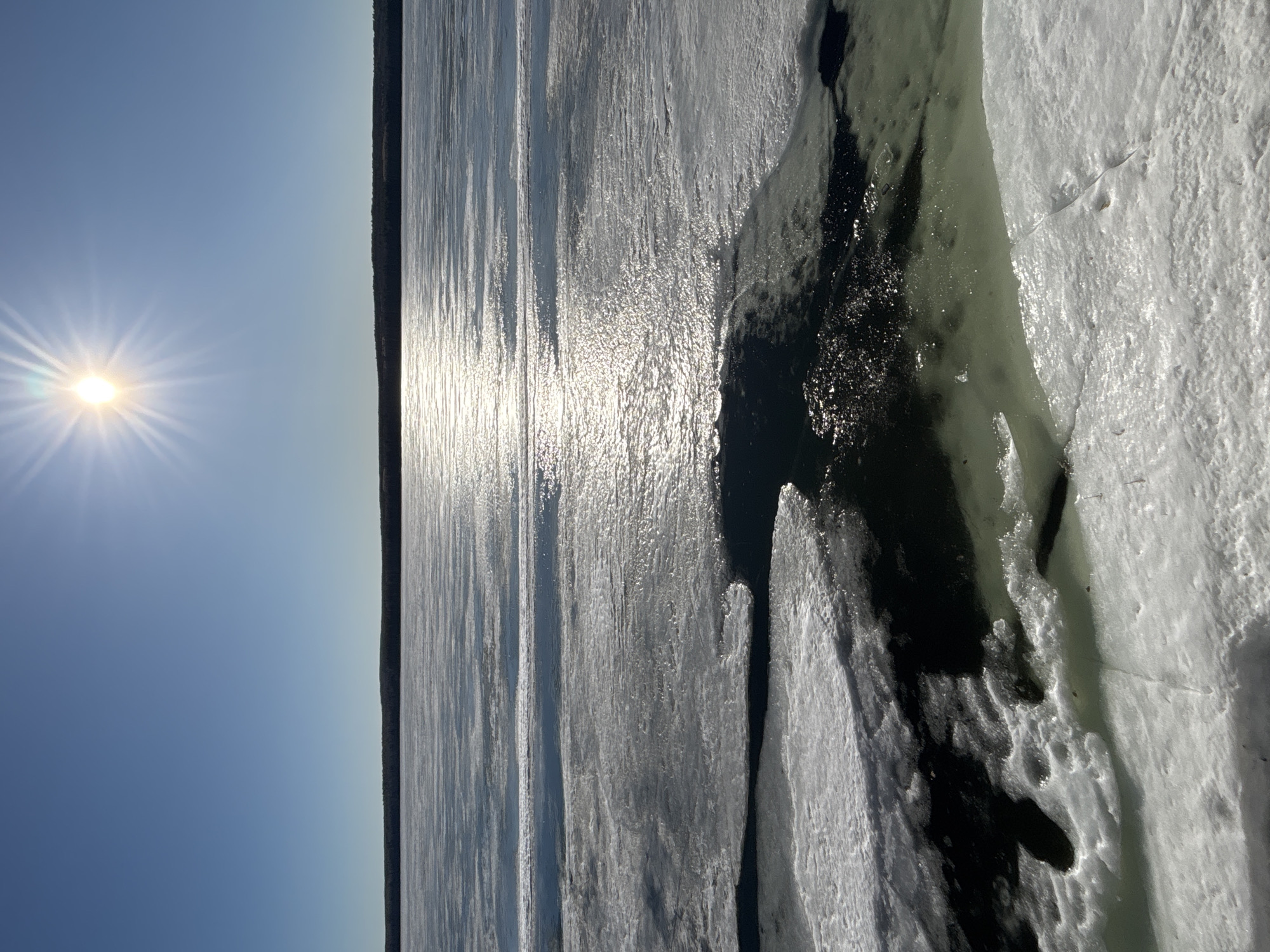 The image shows a frozen river or lake under a clear blue sky with a bright sun. The ice is starting to break apart, with some sections showing open water. A dark tree line can be seen in the distance across the water. The picture appears to be taken at ground level from the edge of the ice. The location is Chaikovsky, Russia.