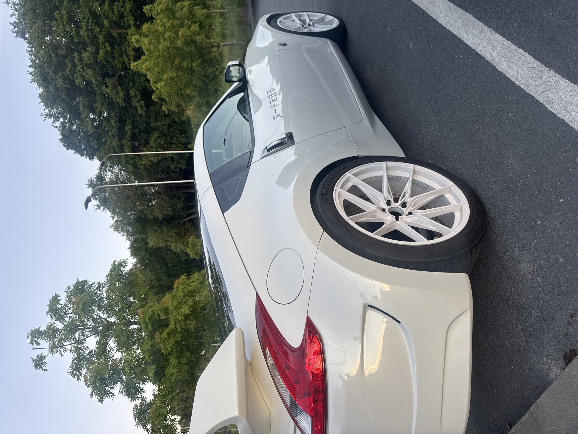 A white Nissan 370Z sports car is parked on asphalt in a lot with a white line marking a parking space. The car has a large white spoiler, custom white rims, and a Japanese decal on the passenger side door. In the background, there are trees and two streetlights. The sky is clear and blue. This image was taken on August 31, 2023, at approximately 8:00 PM in Tallinn, Estonia.