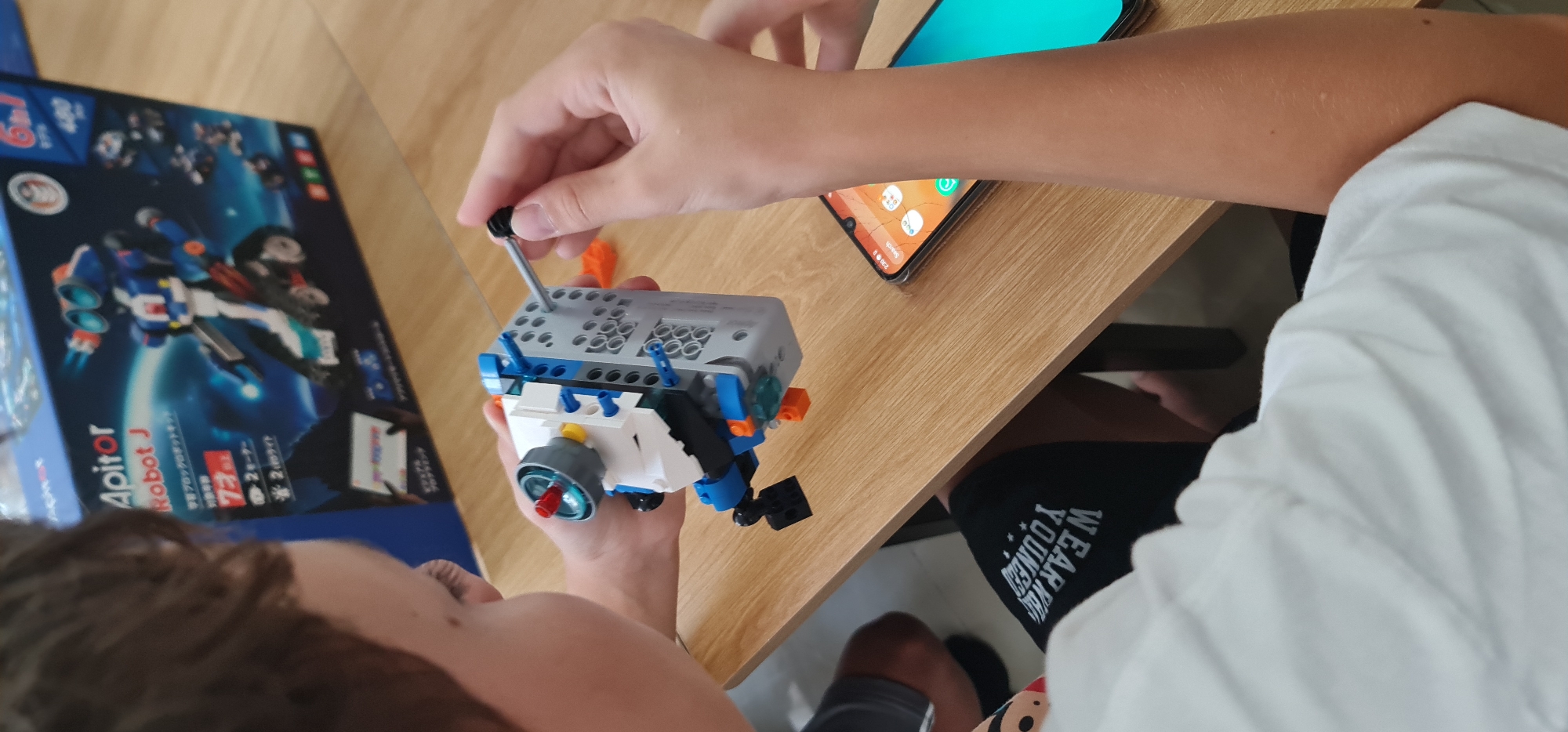 Here's a detailed description of the image:
**Scene:** The image shows a child assembling a robot toy on a wooden table. The environment is likely indoors.
**People:** A child is visible from the shoulders up, focused on assembling the toy. Arms and hands are visible.
**Subjects/Objects:**
* A partly assembled grey, white, blue, and orange robot toy held by the child. A screwdriver is being used to secure it.
* The box for the toy, labeled "Apitor Robot J", is visible in the background.
* A smartphone (partially visible) with a turquoise screen lies on the table.
* The child is wearing black shorts with white writing on the thigh and a white t-shirt.
**Actions/Interactions:** The child is actively building the robot toy.
**Environment:** The scene is well-lit. The wooden table suggests a home environment.
The image does not include any details that would suggest a specific location such as the city of Đà Nẵng, Vietnam.