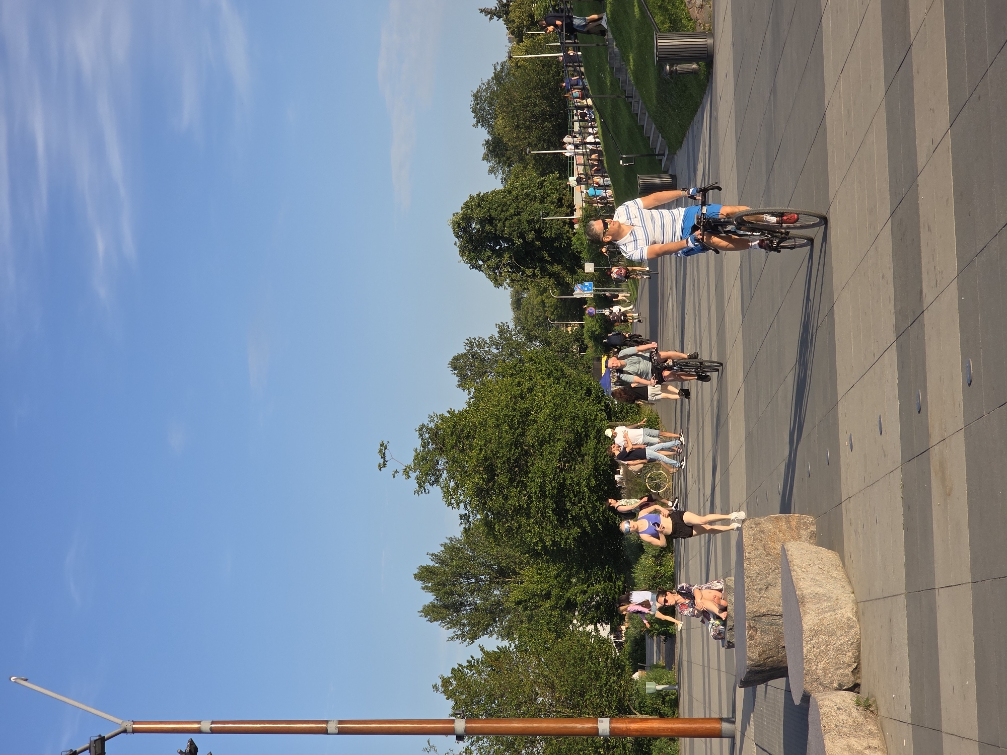 The image depicts a sunny day in Warsaw, Poland, likely in a park or along a waterfront promenade. In the foreground, a man wearing a striped polo shirt and blue shorts is cycling on a paved path. Behind him, another man in a grey t-shirt is also on a bicycle, heading in the opposite direction. To the left, a woman is jogging in athletic wear, and a woman is sitting on a large rock.
Further back, several other individuals are walking and cycling. Trees and green grass line the path and a slope on the right side of the image. A tall wooden lamppost with lights is prominent on the left. The sky is clear blue with a few wispy clouds. The scene suggests a recreational area where people are enjoying outdoor activities.