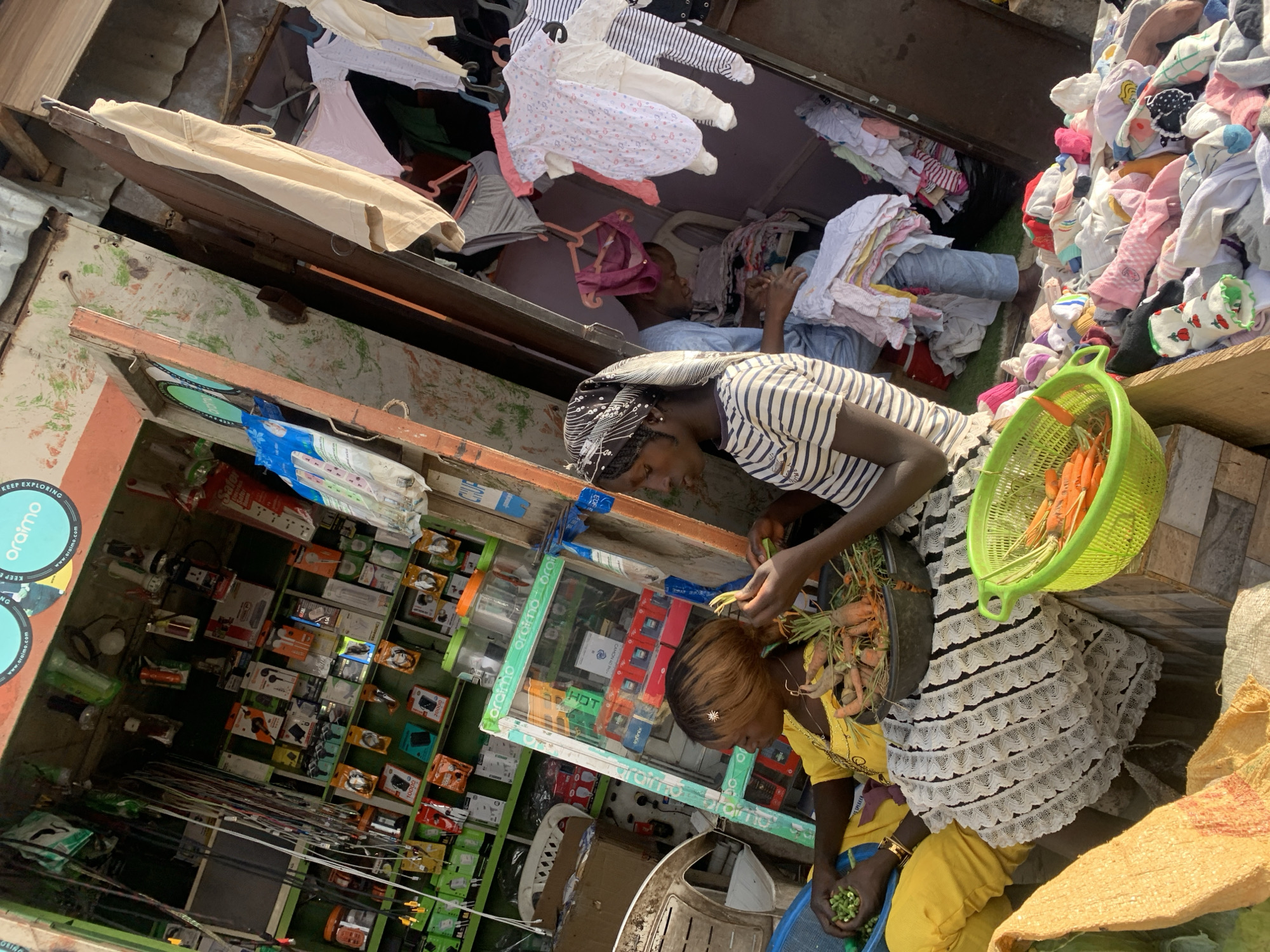 This image captures a vibrant street scene, likely a market or a cluster of storefronts, in Jos, Nigeria, during daylight hours. The perspective is rotated ninety degrees, making the ground appear as a wall on the right.
In the foreground, two women are engaged in preparing produce. The woman on the left, wearing a striped black and white top and a black headscarf, is handling a bunch of carrots with green tops. A bright green plastic basket full of carrots and a dark bowl containing more carrots are placed next to her. The second woman, dressed in a yellow top and skirt, is seated on a low blue stool or container, peeling small green pods, possibly peas or beans. Her left hand wears a golden bracelet.
Behind the women, to their left, is a small electronics shop. Its shelves are packed with various items including phone chargers, speakers, and mobile accessories. Visible text on the products and displays includes brand names like "oraimo" (with a circular sticker also showing "KEEP EXPLORING" and "www.oraimo.com"), and "infinix." Other text fragments like "HOT," "SPEAKER," "CHUR" (likely part of 'CHARGER'), and numbers "10000," "20000" are also discernible.
To the right of the women and extending into the background, there's an area densely filled with clothing. This includes baby clothes, such as onesies and sleepsuits, hanging on hangers and piled up. Another individual can be seen in the background, seemingly organizing or sitting amidst piles of adult and children's clothing. Large, coarse woven sacks, possibly for produce or goods, are visible on the ground to the far right. The overall scene suggests a bustling commercial environment where various goods are sold side-by-side.