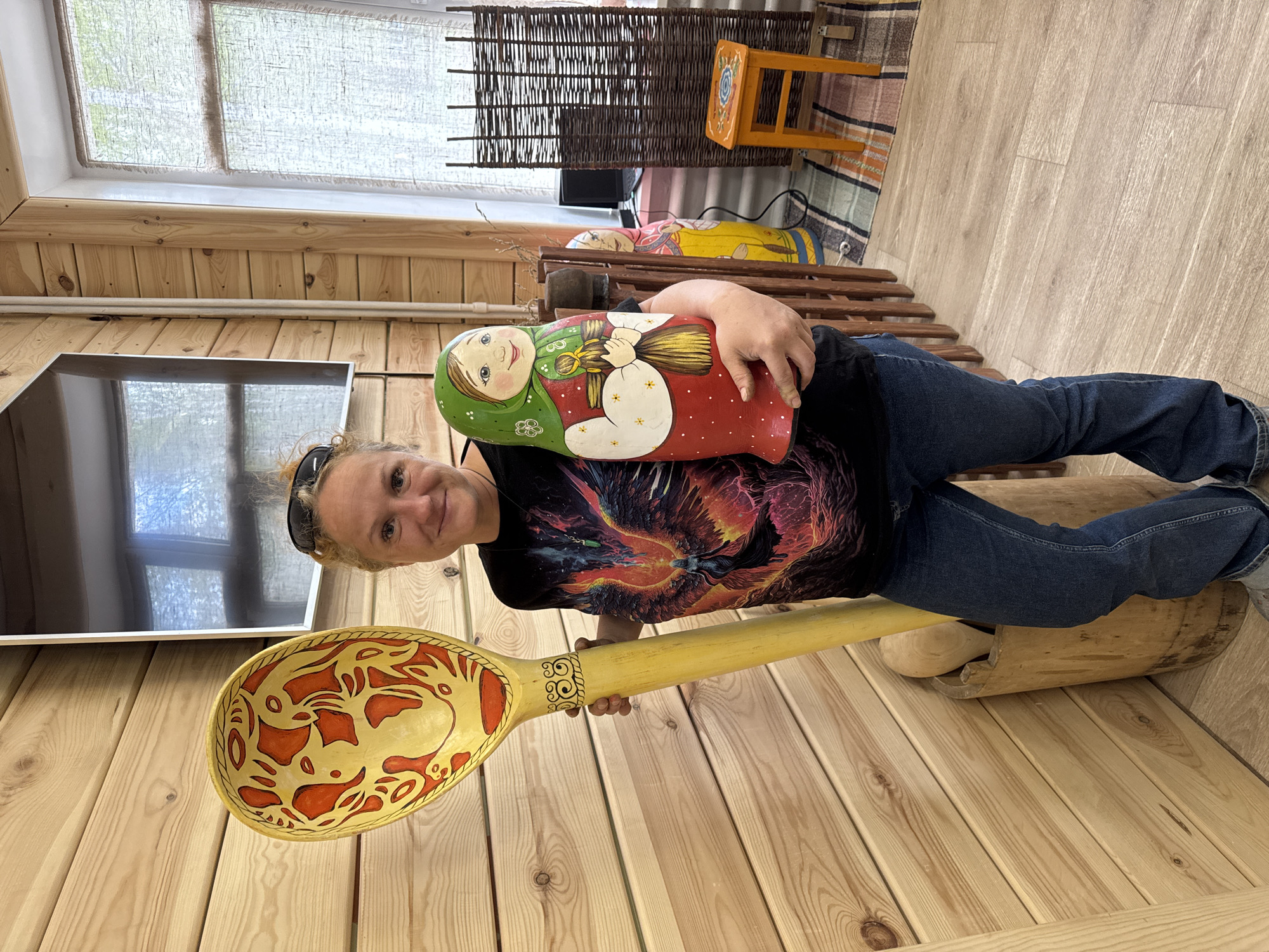 A woman is posing with two Russian-themed objects inside what appears to be a wooden building, likely an attraction in the city of Chaikovsky, Russia. She is holding a large Matryoshka doll and a massive wooden spoon with a decorative pattern. The woman is wearing jeans and a t-shirt with a graphic image, and has sunglasses on her head. The room is paneled with wood, and has a window, a TV, a small yellow stool, a woven room divider, and a bench. There is another Matryoshka doll on the bench. The floor is also made of wood.