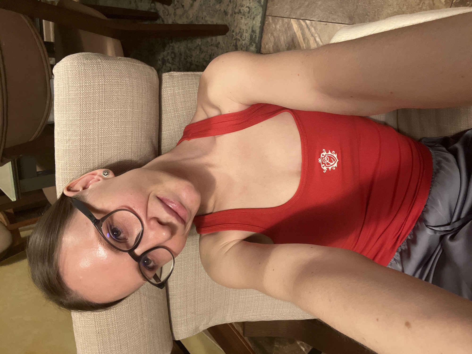 A person with glasses and a red tank top is lying on a light-colored upholstered chair. The person is facing the camera. Part of a wooden chair is visible on the left. The floor is tiled. The location is not specified.