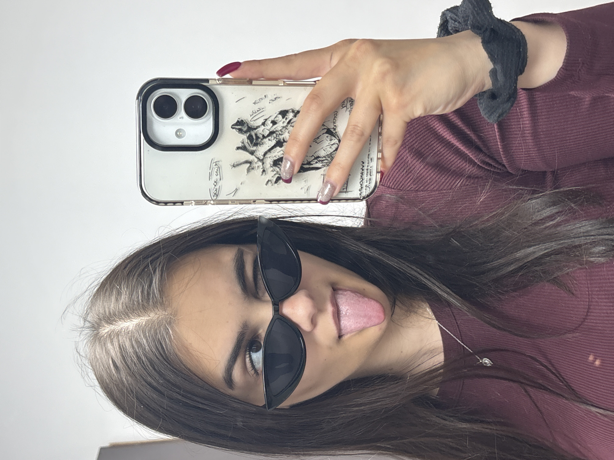 A young woman is posing for a selfie while sticking out her tongue. She is wearing sunglasses and a burgundy shirt. Her long dark hair is visible. She is holding a white smartphone with a clear case featuring a sketch design, with a burgundy manicure visible on her nails. On her wrist, she has a black scrunchie. A thin silver necklace is visible around her neck. The background is a plain white wall. The location is unknown; there is no information in the image to suggest it is in Blagoevgrad, Bulgaria.