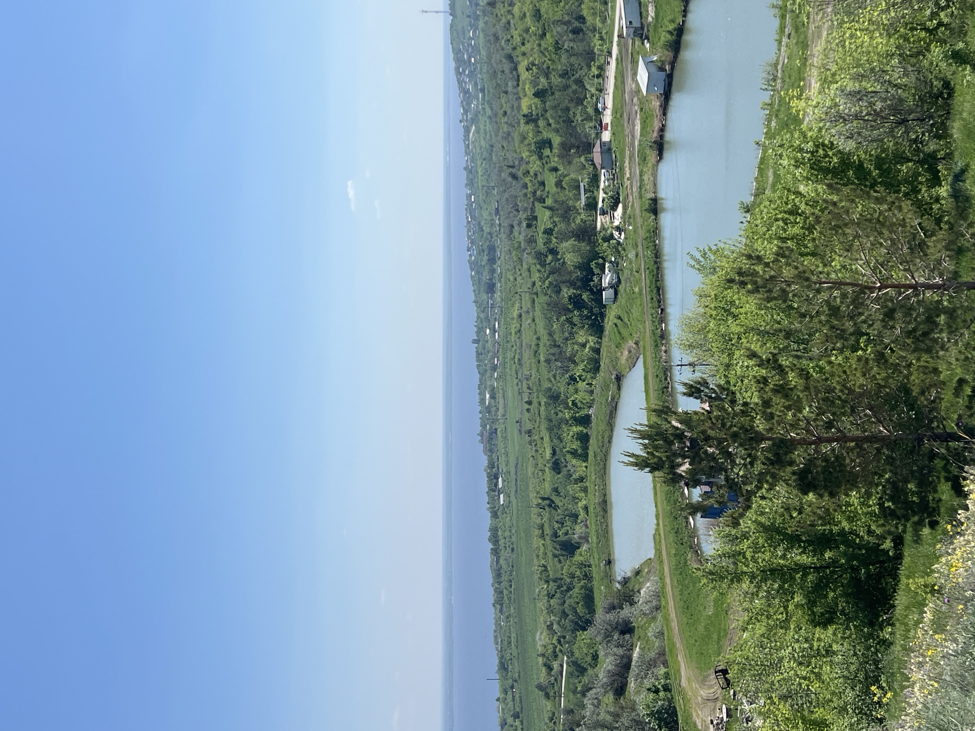 The image shows a view of Khvalynsk, Russia. In the foreground are green trees and bushes. Beyond that is a canal with light blue water. A road runs alongside the canal, and some small buildings can be seen next to the road. In the distance are fields and the Volga River. The sky is clear and blue.