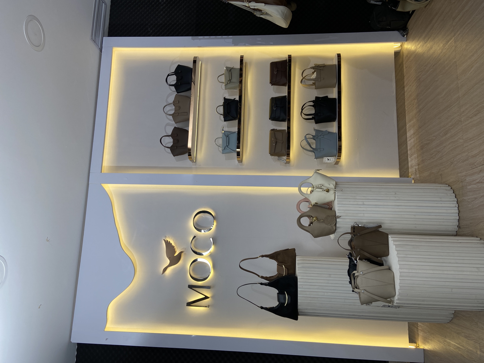 The image shows a display of handbags in what appears to be a store, possibly in North Jakarta, Indonesia. The handbags are arranged on white shelving units with decorative lighting. There are three rows of smaller handbags displayed against a flat white surface with lighted trim. Below, the "MOOCO" logo in gold with a bird emblem is seen. There are also shelves made from a corrugated material displaying more bags of varied style and color. The flooring is a light shade, possibly tile.