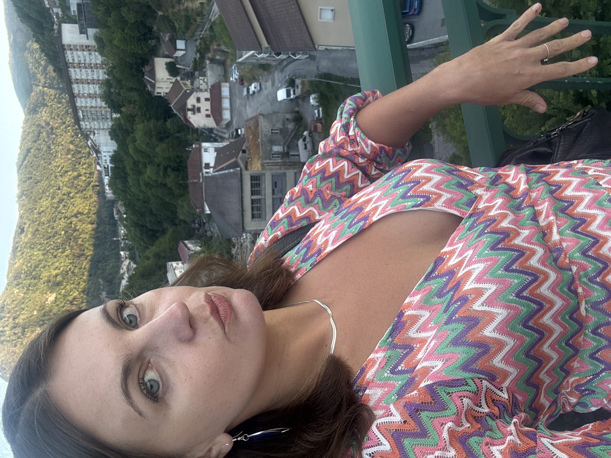 The image shows a woman standing on a bridge, looking at the camera. She has brown hair and blue eyes, and she is wearing a colorful zigzag-patterned dress. Her left hand is resting on a green railing, and she is wearing a ring on her left ring finger. In the background, there are buildings and trees on a hillside. The city of Saint-Claude, France, is known for its beautiful natural surroundings and historic architecture. This image appears to capture a scenic view within the city.