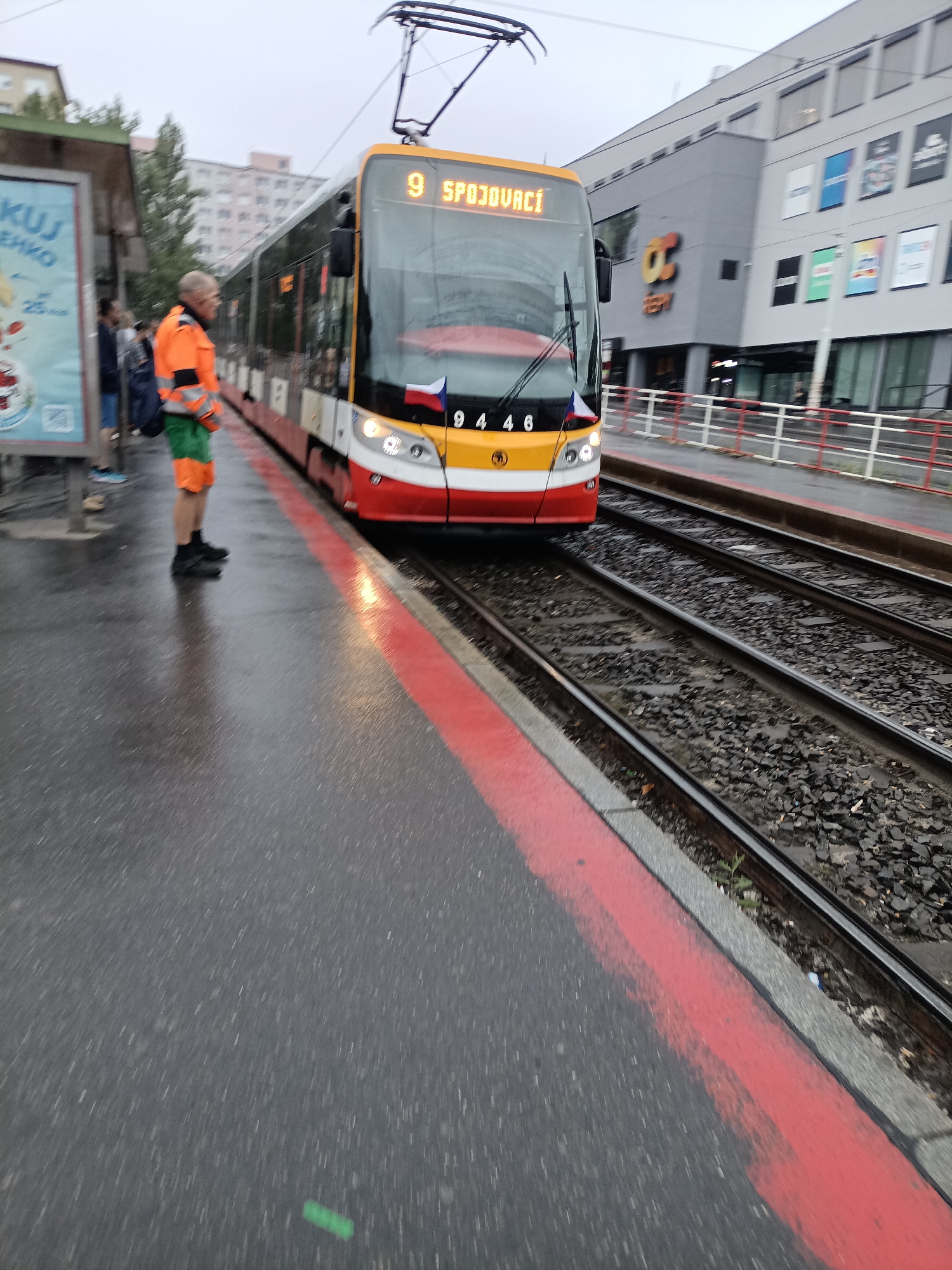 A yellow and red tram is stopped at a tram station on a rainy day in Prague, Czechia. The tram has the number "9" displayed on its front, along with the destination "SPOJOVACÍ". A man wearing an orange high-visibility vest and green pants stands on the platform, looking towards the tram. The platform is wet, indicating recent rain, and a red line is painted along the edge. In the background, a modern building with shops and a large "OC" logo is visible. Several people are standing on the platform waiting for the tram, and a billboard advertising something with strawberries is to the left. The tram tracks are visible next to the platform.
