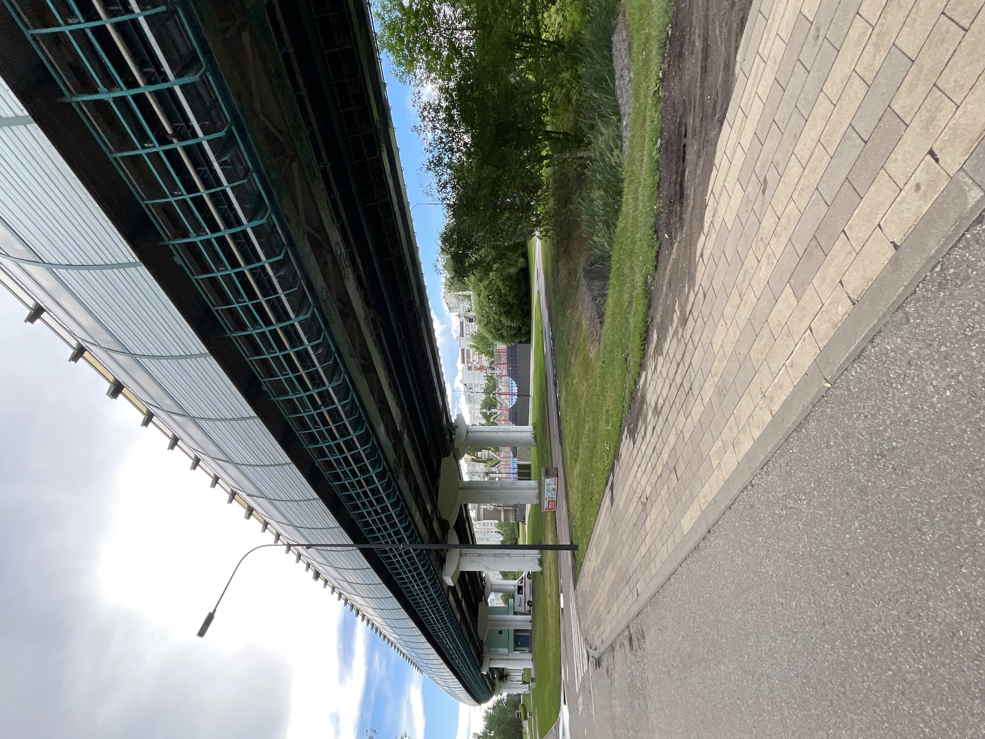 The image depicts a view of an overpass and its underside, situated in Moscow, Russia. The overpass structure is prominent, with its supporting pillars and a metal grid visible underneath. To the right of the overpass, there is a paved pedestrian walkway made of light-colored rectangular tiles, bordered by a strip of grass and then a road. Beyond the grass, trees and buildings can be seen in the background. The sky above is partly cloudy, suggesting daytime. There is a street lamp attached to the underside of the overpass. No people are visible in the image. The scene appears to be a public urban space, likely a park or a pathway alongside a road and under a bridge. There is some text on a red and white sign visible near the walkway.