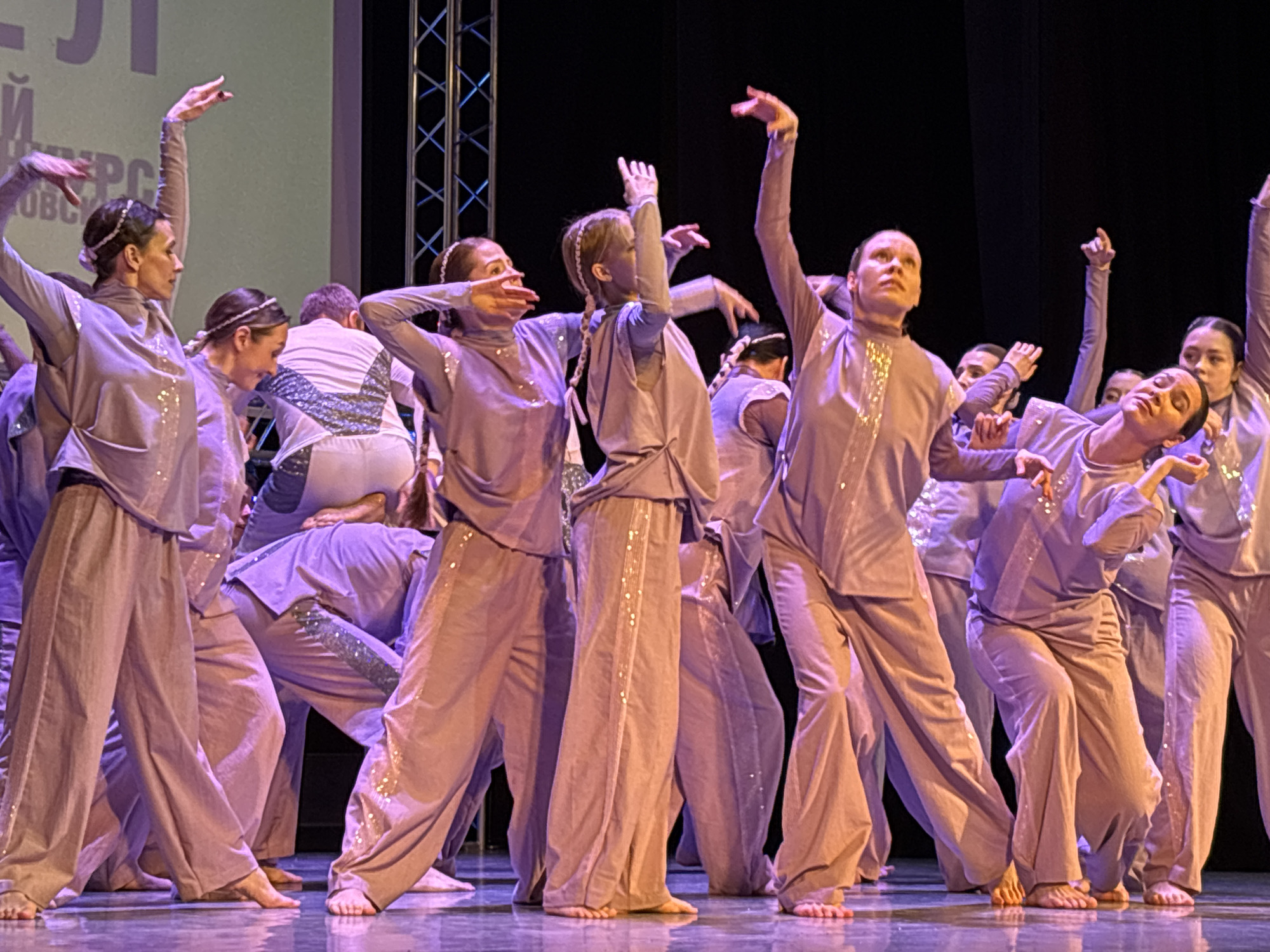 A group of barefoot dancers in matching light purple costumes with silver accents are performing on a stage. The costumes consist of long-sleeved shirts and loose pants. The dancers are arranged in a dynamic composition, with some standing tall and others bending or crouching. Their arms are raised and positioned in various expressive poses. The stage has a shiny surface that reflects the light, and there is a dark background behind the dancers, with metal support structures visible on one side. The location is likely a theatre or performance hall in the city of Chaikovsky, Russia, based on the Cyrillic text visible on a screen in the background.