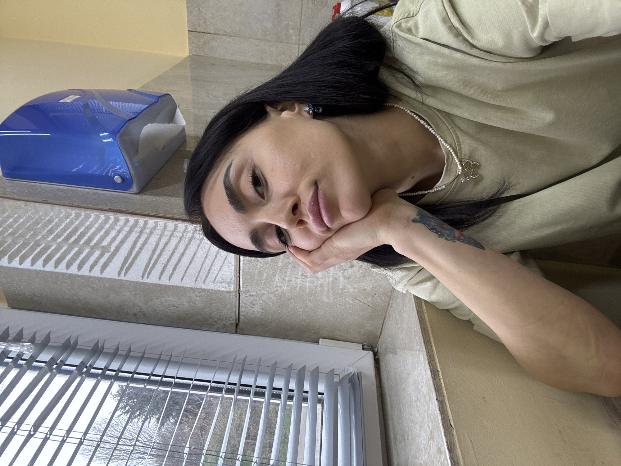 The image displays a close-up of a female individual in an indoor setting. The subject has dark hair, styled with a part on the left side, and is wearing a light olive-green crew-neck shirt, a white pearl-beaded necklace with a gold-colored butterfly pendant, and dark blue or black drop earrings. A colorful tattoo is partially visible on her upper left arm. Her right hand supports her chin, with fingers resting on her cheek and her thumb near her jawline. Her gaze is directed towards the camera with a neutral expression and slightly pursed lips.
To the immediate left of the individual is a white-framed window fitted with horizontal white venetian blinds, partially open. Through the blinds, an exterior view reveals bare trees under an overcast sky. Below the window is a light-colored, possibly tiled, window sill. The interior wall adjacent to the window is covered with light beige, textured rectangular tiles. Further to the right and above the subject's head, a blue wall-mounted dispenser, consistent with a paper towel or hand dryer unit, is affixed to a yellowish-beige painted wall, which also displays diagonal shadows from the window blinds. The lighting appears to be a mix of natural light from the window and interior artificial light. The specific location within the city of Tbilisi, Georgia, cannot be determined from the visual information provided.
