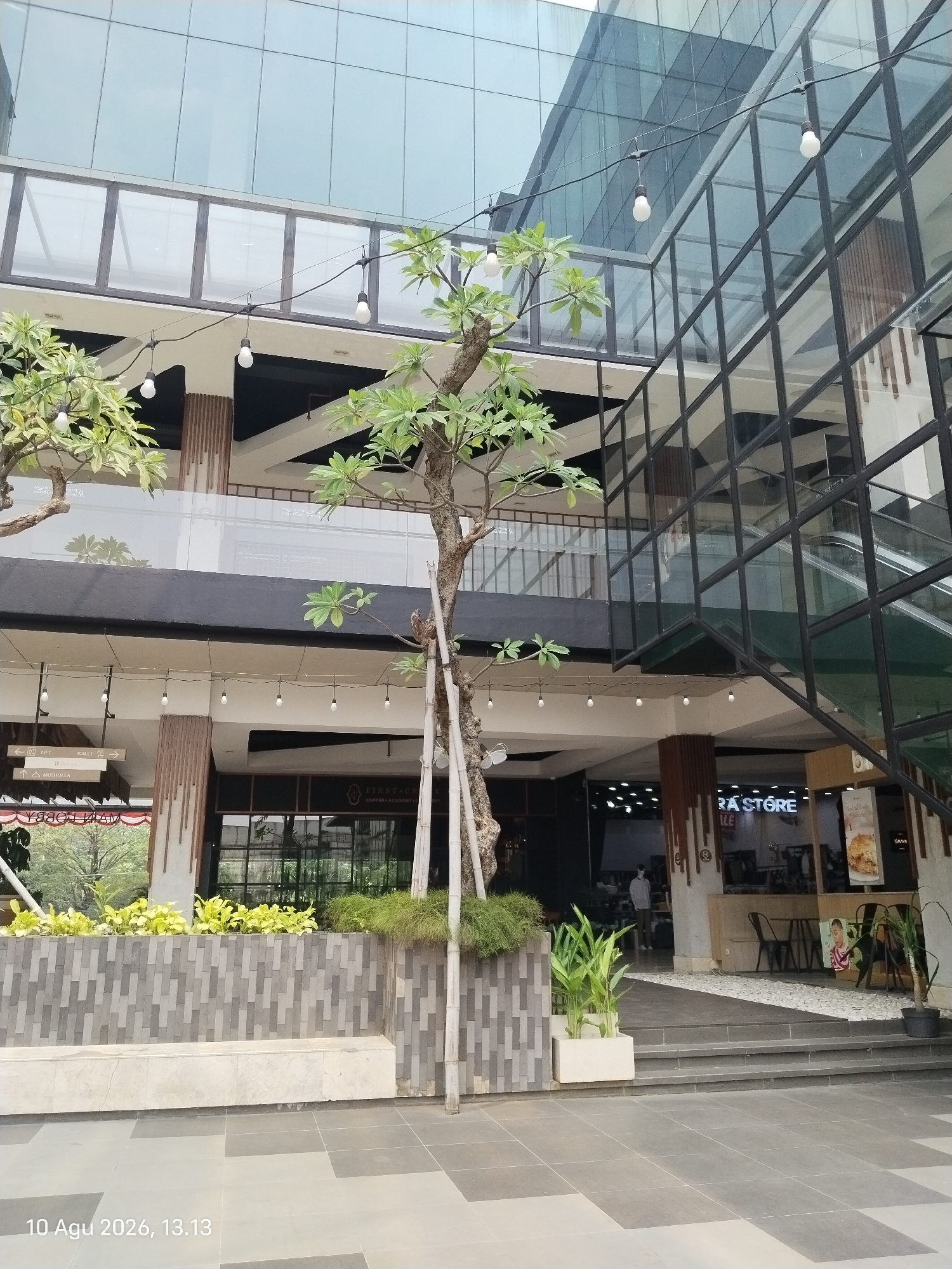 The image depicts the exterior of a modern multi-story building, likely a shopping mall or office complex, located in East Jakarta, Indonesia. The architecture features extensive use of glass and metal, with large glass facades reflecting the sky and surrounding environment. String lights with exposed bulbs are hung across several levels of the building, suggesting an outdoor or semi-outdoor common area.
In the foreground, a paved walkway with a grey and dark grey tiled pattern leads towards the building's entrance. A low, grey tiled wall with a light-colored stone bench runs along the walkway, adorned with potted plants, including vibrant green leafy plants and tall, slender grass-like foliage. A young tree with thin trunks and sparse green leaves stands near the right side of the frame, its trunk supported by bamboo stakes.
The building's lower levels reveal shop fronts and entrances. One sign reads "RA STORE". Another visible sign indicates "FIRST CH..." likely for a coffee shop or academy. An escalator is visible through the glass facade on the right side, leading to an upper level. Several people are partially visible inside the building, including one person standing near a shop entrance and another person walking on an upper walkway. The overall impression is of a commercial and public space designed with contemporary aesthetics. The timestamp in the lower-left corner reads "10 Agu 2026, 13.13".