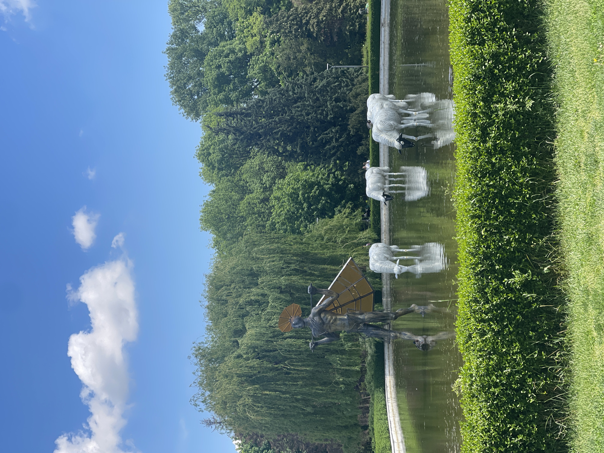 The photograph captures a tranquil scene in a park, featuring a man-made pond or water feature. The pond is adorned with sculptures: one depicting a man with a parasol and what appears to be wings, and another set depicting several sheep. The statues are positioned in the water, creating reflections on the water's surface. The water is surrounded by manicured greenery, including a neatly trimmed hedge on one side and a lush green lawn on the other. Tall, mature trees form the backdrop, providing shade and adding to the natural ambiance of the setting. The sky is blue with some white clouds.
Based on the style of the sculptures, the park setting, and visual landmarks, the location appears to be in Sofia, Bulgaria. Specifically, the sculptures are characteristic of those found in Borisova Gradina (Boris' Garden), a large park in the city. The man-with-parasol sculpture is known as the "Aviator" sculpture.