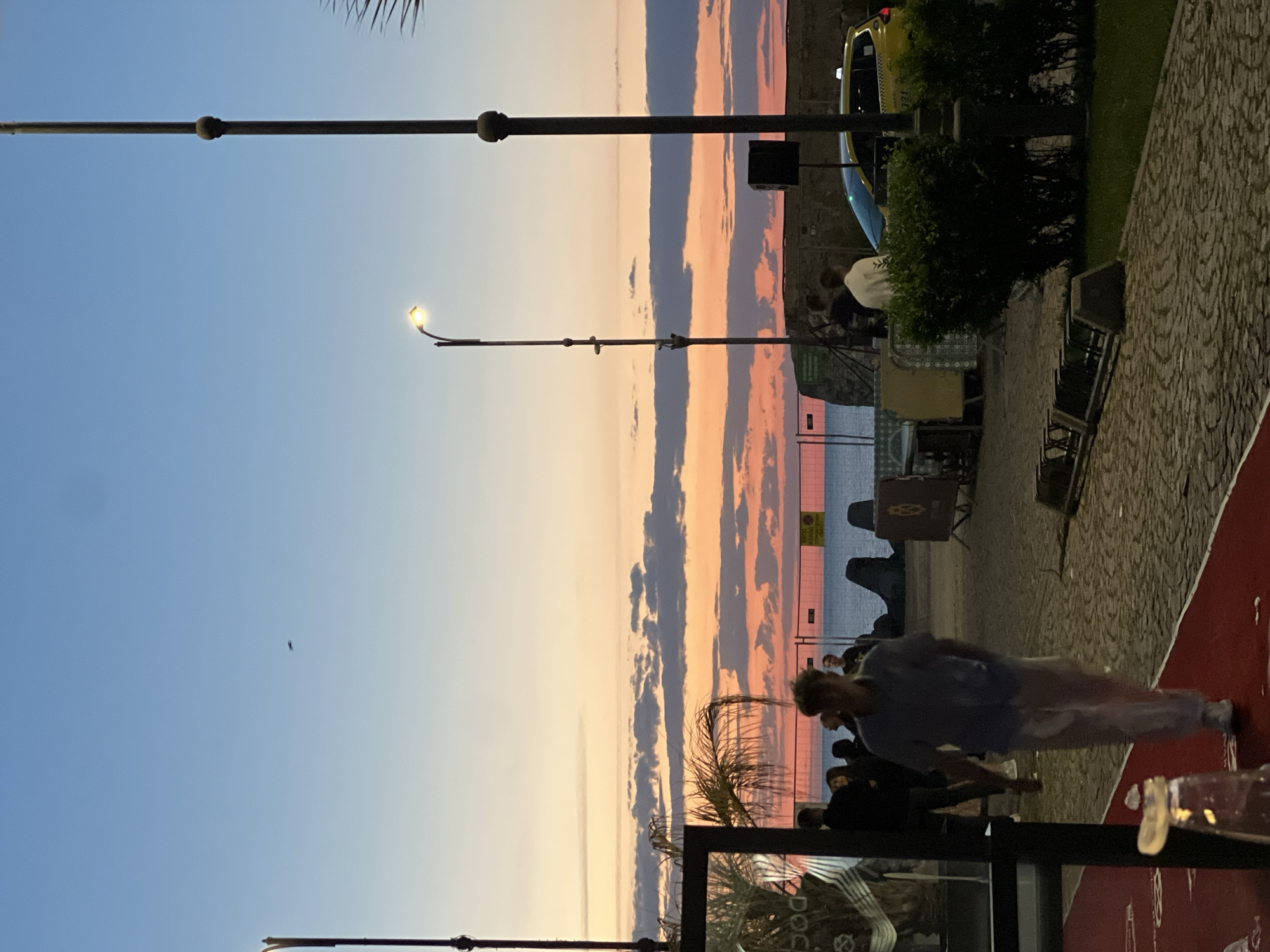 The image depicts an outdoor scene along a promenade during sunset, likely in a coastal area. The sky is a gradient of blue and orange, transitioning to pink hues at the horizon, with streaky clouds present. A street lamp stands prominently in the middle-ground against the sky.
People are present in the lower half of the image; some are sitting at tables and others are walking on a red-surfaced promenade. There's outdoor seating with tables and chairs; some tables appear to be occupied. Green bushes and trees are also present. A part of a yellow taxi cab is visible in the background.
Given the context of Burgas, Bulgaria, it is plausible that the location is near the sea garden or along the coastline of the city.