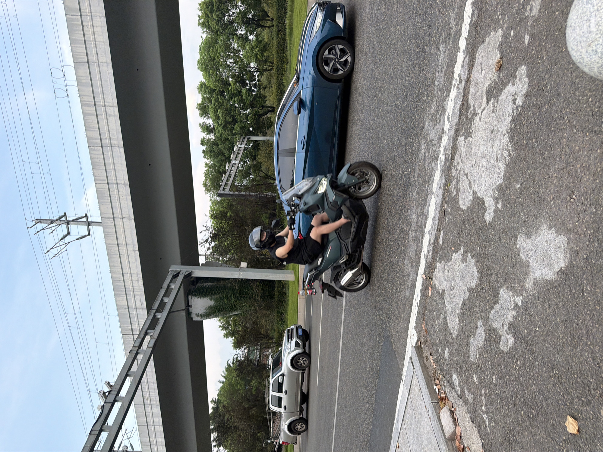 A scooter with a rider wearing a helmet and a black sleeveless shirt and black shorts is in the foreground, on a paved road. The scooter is dark green. To the left of the scooter, a silver pickup truck is driving on the same road. To the right of the scooter, a blue sedan is also driving on the road. In the background, there is a large overpass with overhead power lines and vegetation growing on its support pillars. The sky is overcast. The road has a white lane marker. The foreground shows the edge of the road with a sidewalk and some fallen leaves. The scene appears to be in Jiangjin District, Chongqing, China.