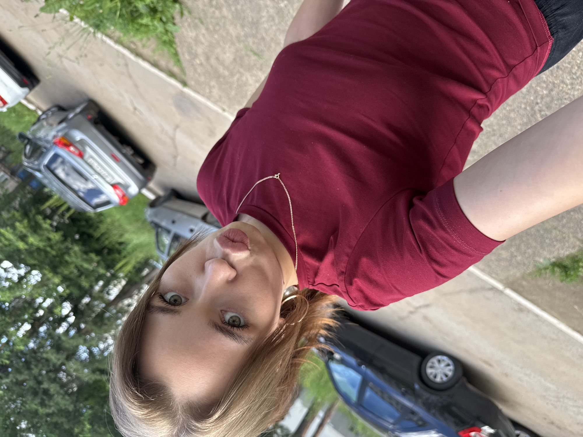 A person is captured in a selfie, with their face taking up a significant portion of the frame. The person has light brown hair, green eyes, and is wearing a maroon t-shirt and a delicate gold chain necklace. Their lips are pursed, creating a playful expression. The background suggests an outdoor setting, possibly a parking lot, with cars visible. To the left, there are trees and greenery, indicating it might be daytime. The overall mood of the image is casual and candid. No visible text is present in the image.