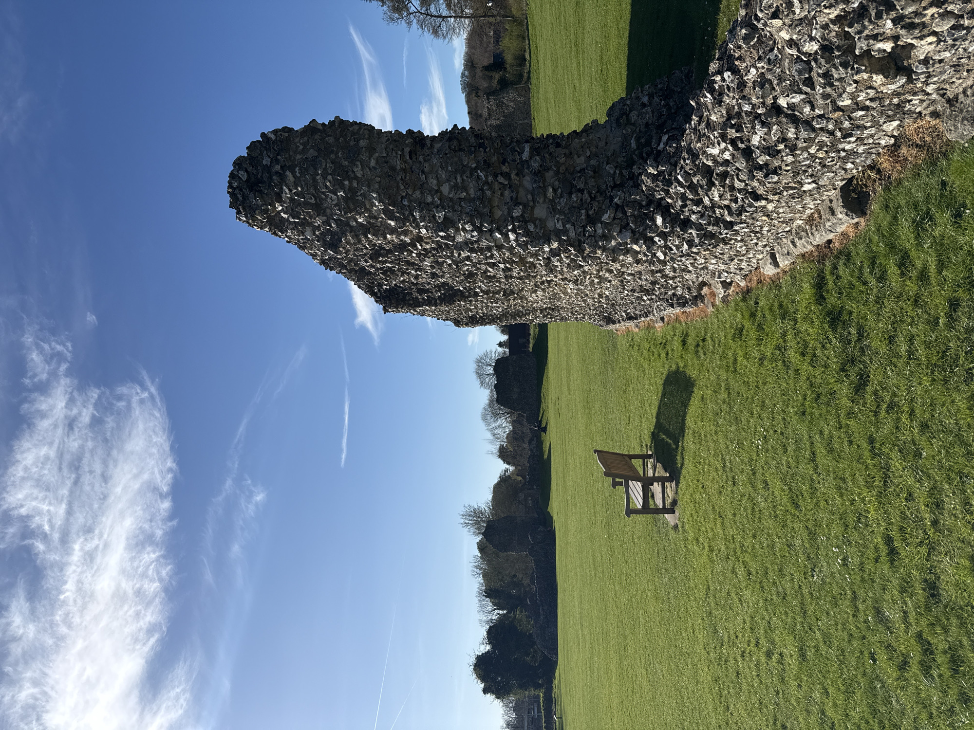 An exterior shot, taken during the day, shows a stone wall, a bench, and a green field, situated at Berkhamsted Castle, a historic site in Dacorum, United Kingdom.
The rough textured, light-gray stone wall runs horizontally across the frame, partially obscuring the landscape behind it. To the right of the wall, a vibrant green field extends outward. A wooden bench, casting a sharp shadow, is positioned on the field, adjacent to the wall. In the background, the tops of trees are visible, as well as a sliver of blue sky with streaks of clouds.