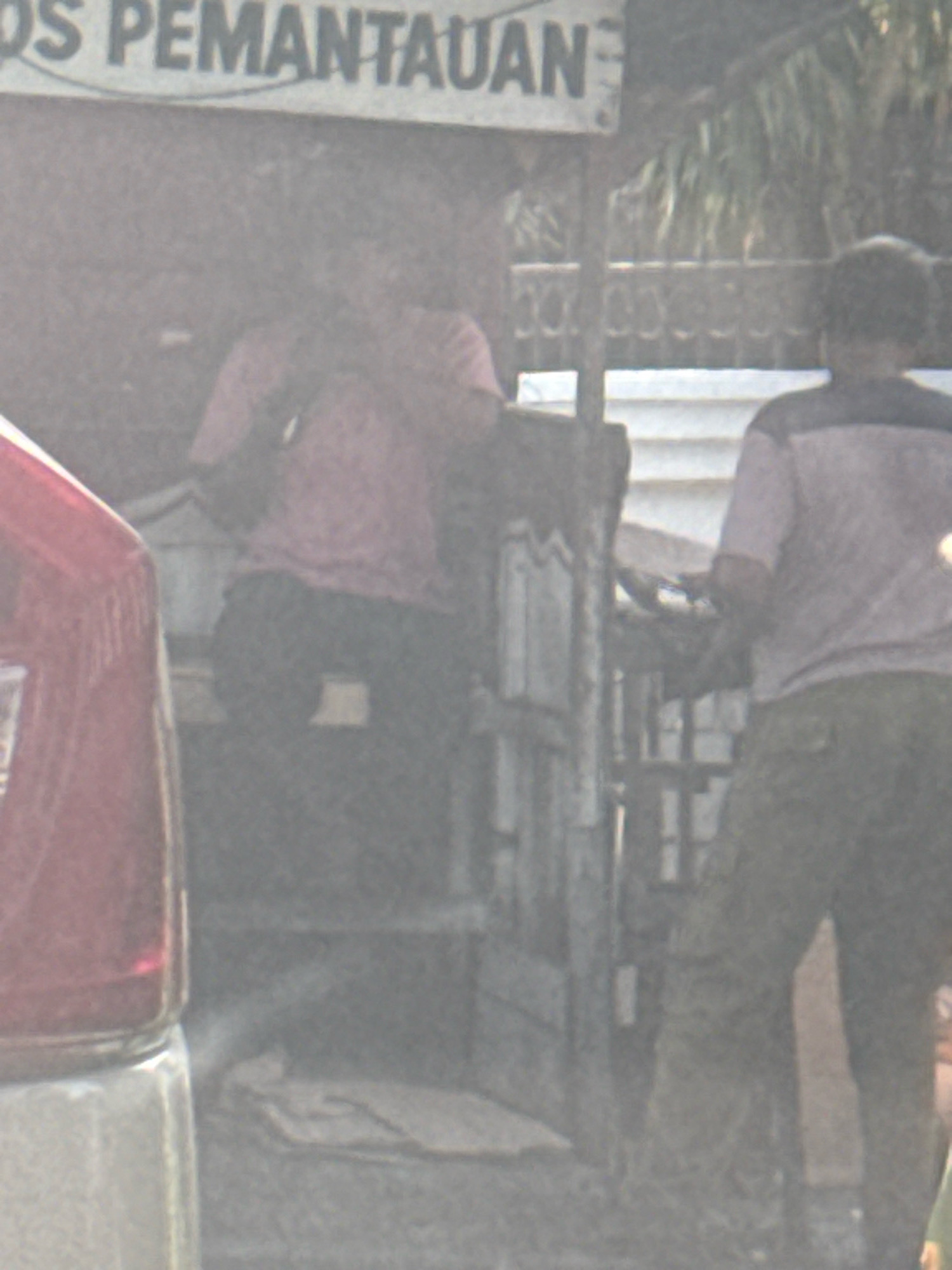 The image shows a blurry outdoor scene with two individuals. A sign with text, partially visible and appearing to say "POS PEMANTAUAN", is mounted on a structure. One person is seated, wearing a pink shirt and dark pants, with their left arm resting on their lap. The other person is standing, wearing a light-colored shirt with a dark collar and dark pants. They are facing away from the camera and appear to be holding something. The environment includes wooden structures and foliage in the background, suggesting an urban or semi-urban setting in Surabaya, Indonesia. The overall image quality is low, with significant blur and low contrast, making precise details difficult to discern.