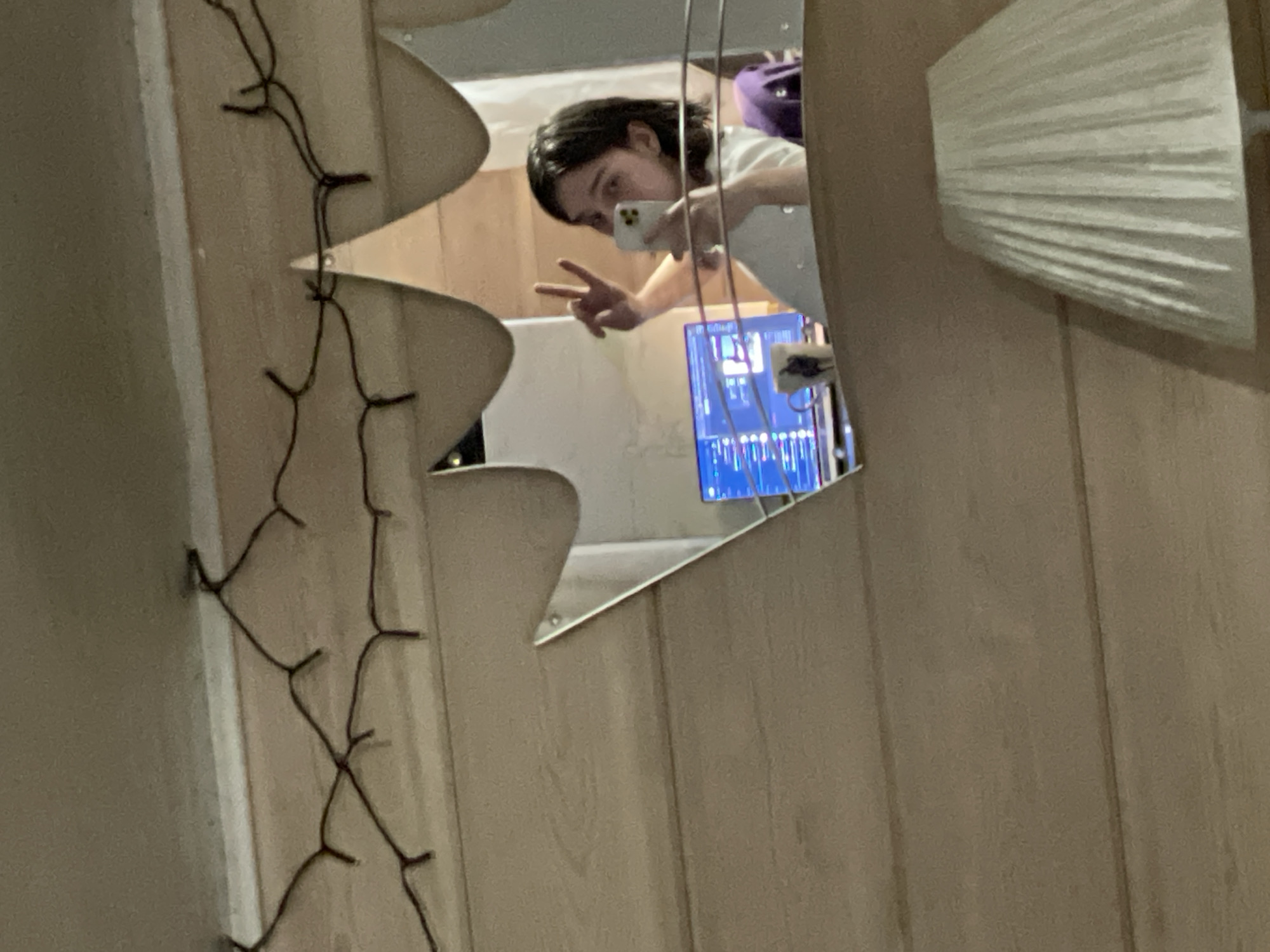 A young woman with dark hair is taking a selfie in a mirror. She is holding a smartphone in her right hand and making a peace sign with her left hand. She is wearing a white shirt. In the background, a computer monitor displays code, a mug sits on a desk, and a purple object is visible. The mirror has a decorative, leaf-like shape. The scene appears to be indoors, with wooden paneling on the wall and a string of lights across the ceiling. The image is cropped to focus on the woman and the mirror's reflection. There is no discernible information about the location in Angarskie Khutora, Russia.