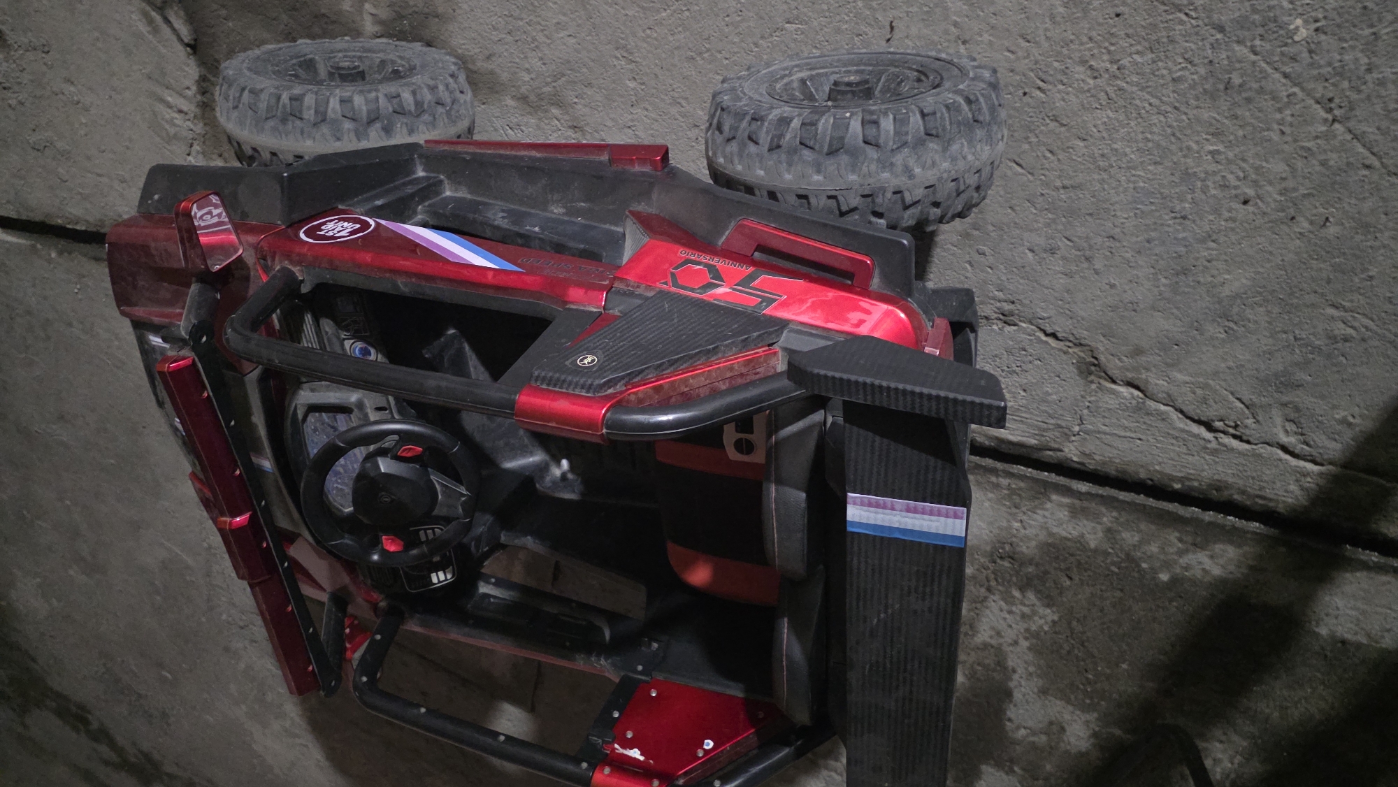 An overhead, slightly tilted, eye-level shot captures a red and black toy ride-on vehicle situated on a cracked concrete surface. The vehicle is a scaled-down replica of an off-road buggy, complete with a steering wheel and what appear to be seats within its open-top interior. Prominently displayed on the side of the vehicle is a "50 ANNIVERSARIO" decal, suggesting it might be a special edition model. The environment appears to be an outdoor space, possibly a garage or an alleyway, with the concrete showing signs of wear and tear. The lighting is dim, casting shadows and creating a somewhat stark atmosphere. There are no people or animals visible in the image. The location is identified as Chuy, Kyrgyzstan.